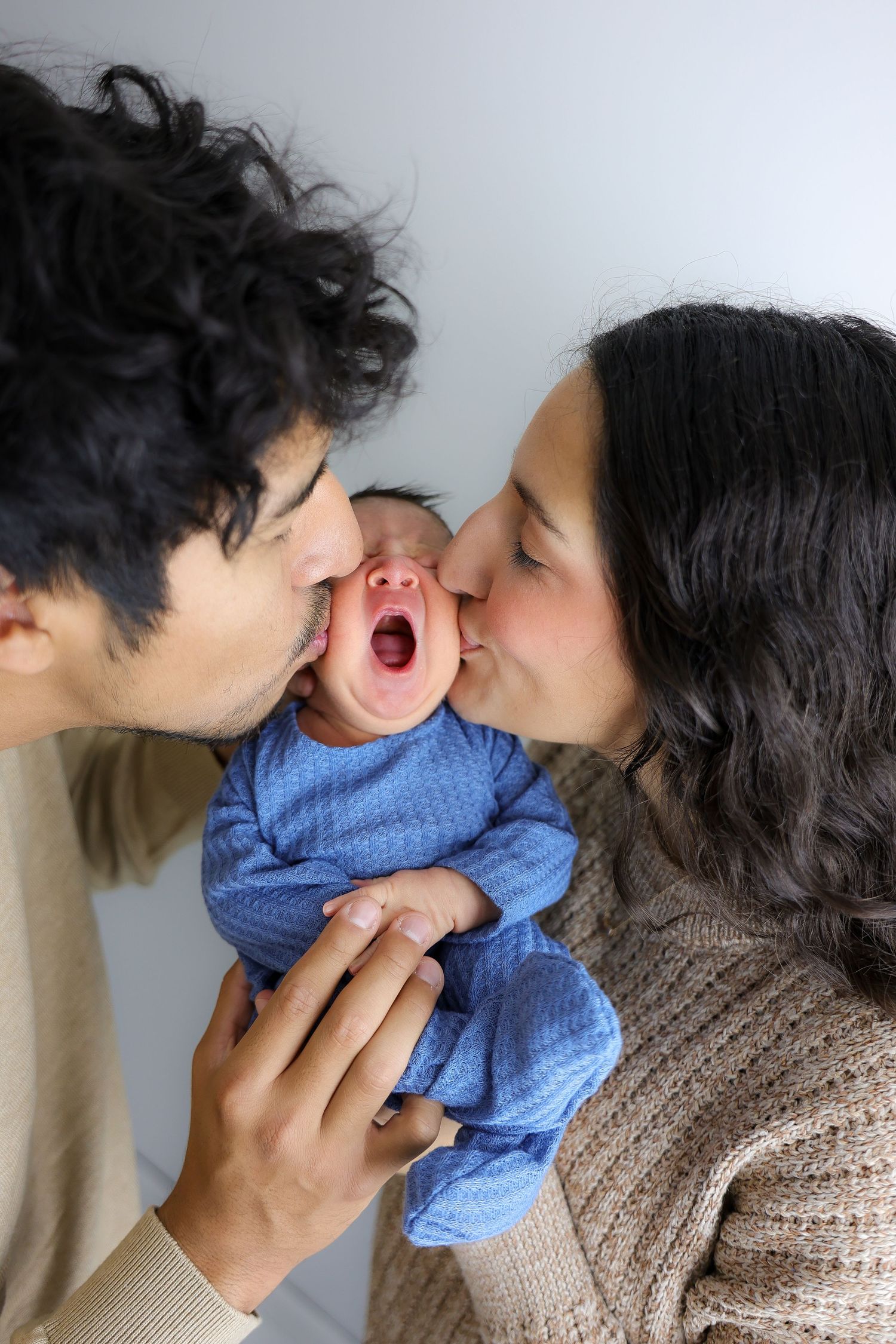 A tender family moment shows parents sharing affectionate kisses with their baby wearing a blue sweater.