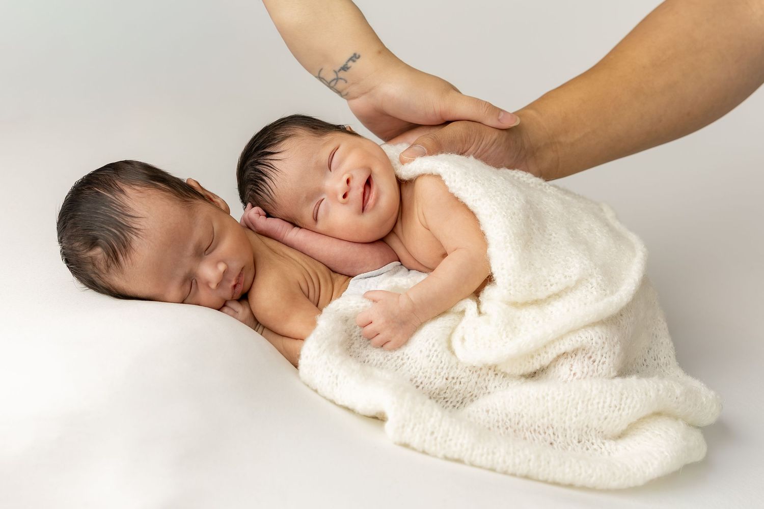 Two sleeping newborn twins wrapped in a soft white blanket, cradled gently by a parent's hands.
