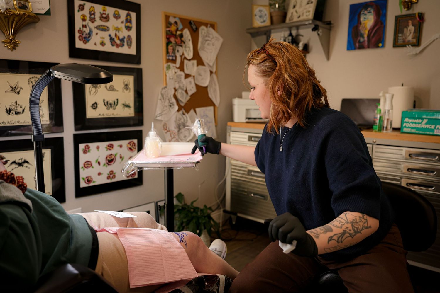 A tattoo artist with red hair and tattooed arms works on a client in a decorated studio filled with framed flash art.
