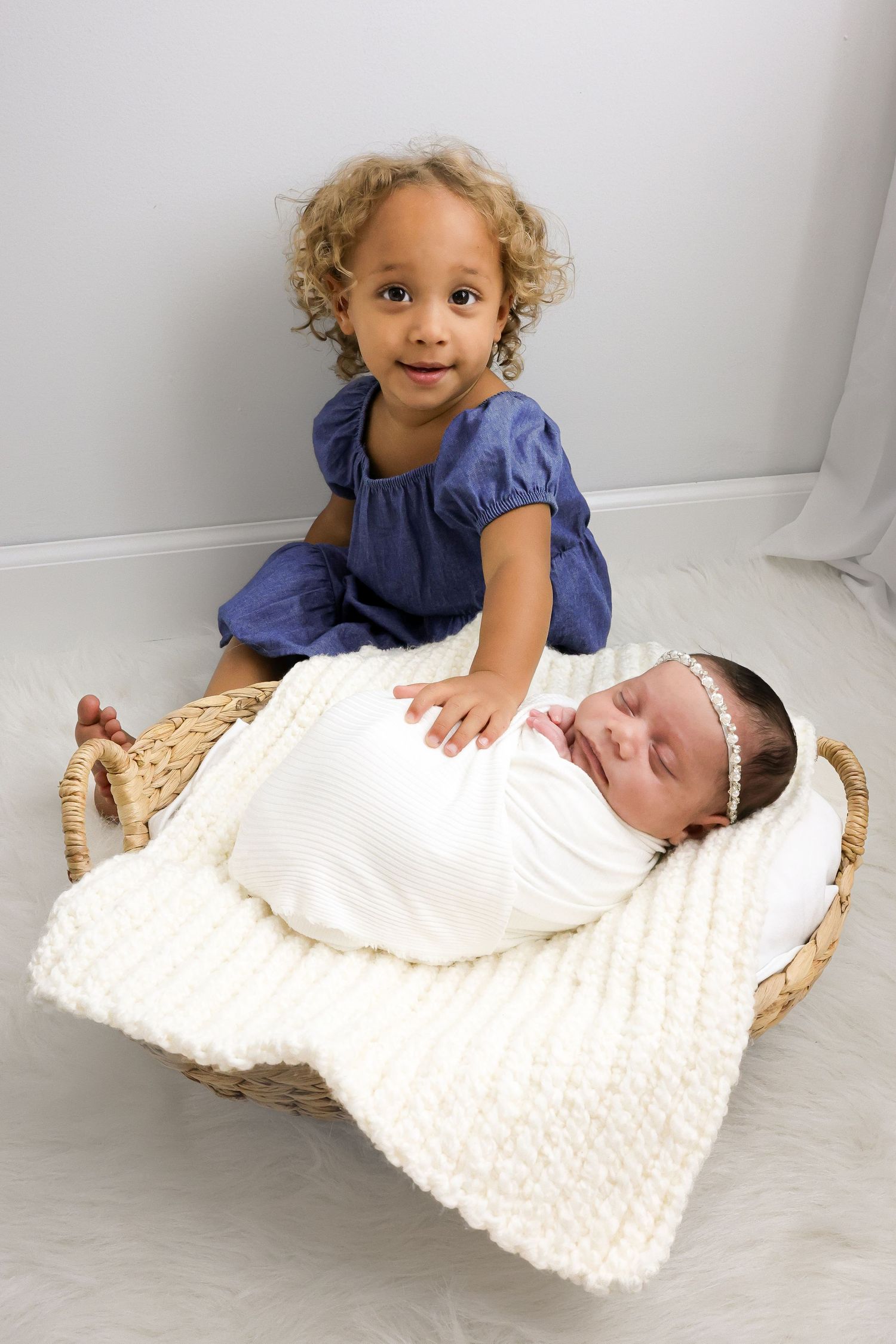 A toddler in a blue dress sits next to a sleeping newborn baby swaddled in white on a woven basket.
