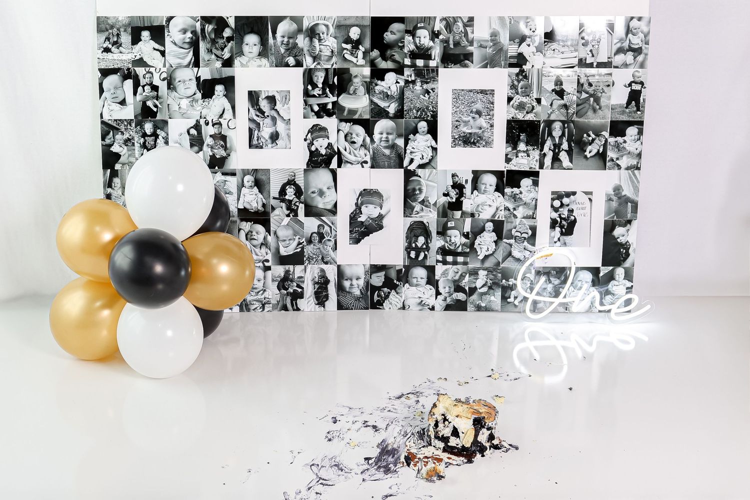 A photo backdrop with black and white photos, gold and black balloons, and cake smash remnants.