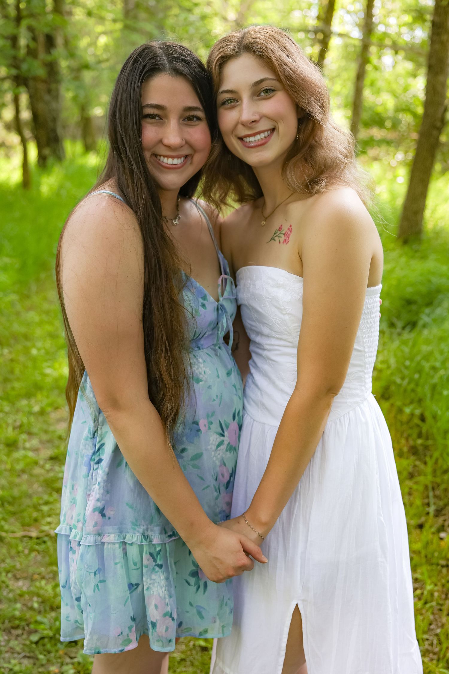 Two friends embrace while wearing floral and white dresses in a wooded outdoor setting.