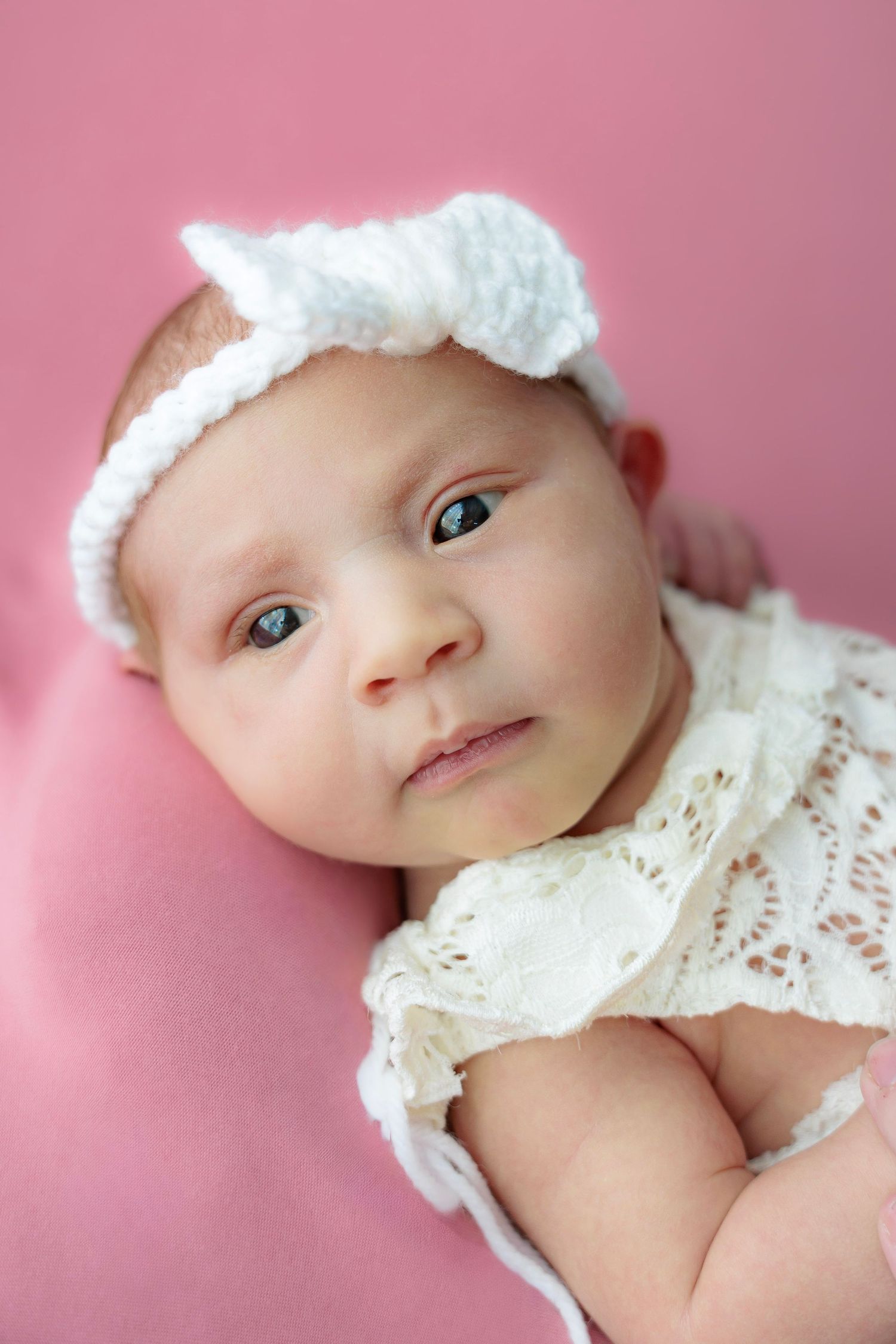 A baby wearing a white lace outfit lying on pink bedding looking at the camera with bright eyes.