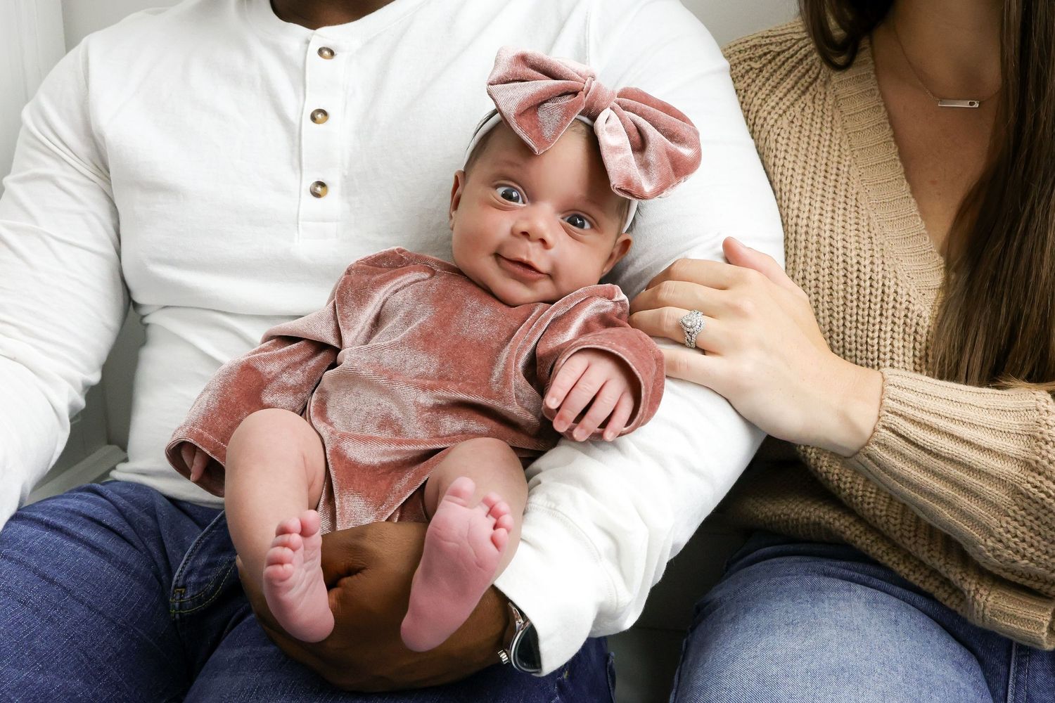 Baby in a white onesie lies on a couch while being held by someone in a beige sweater.