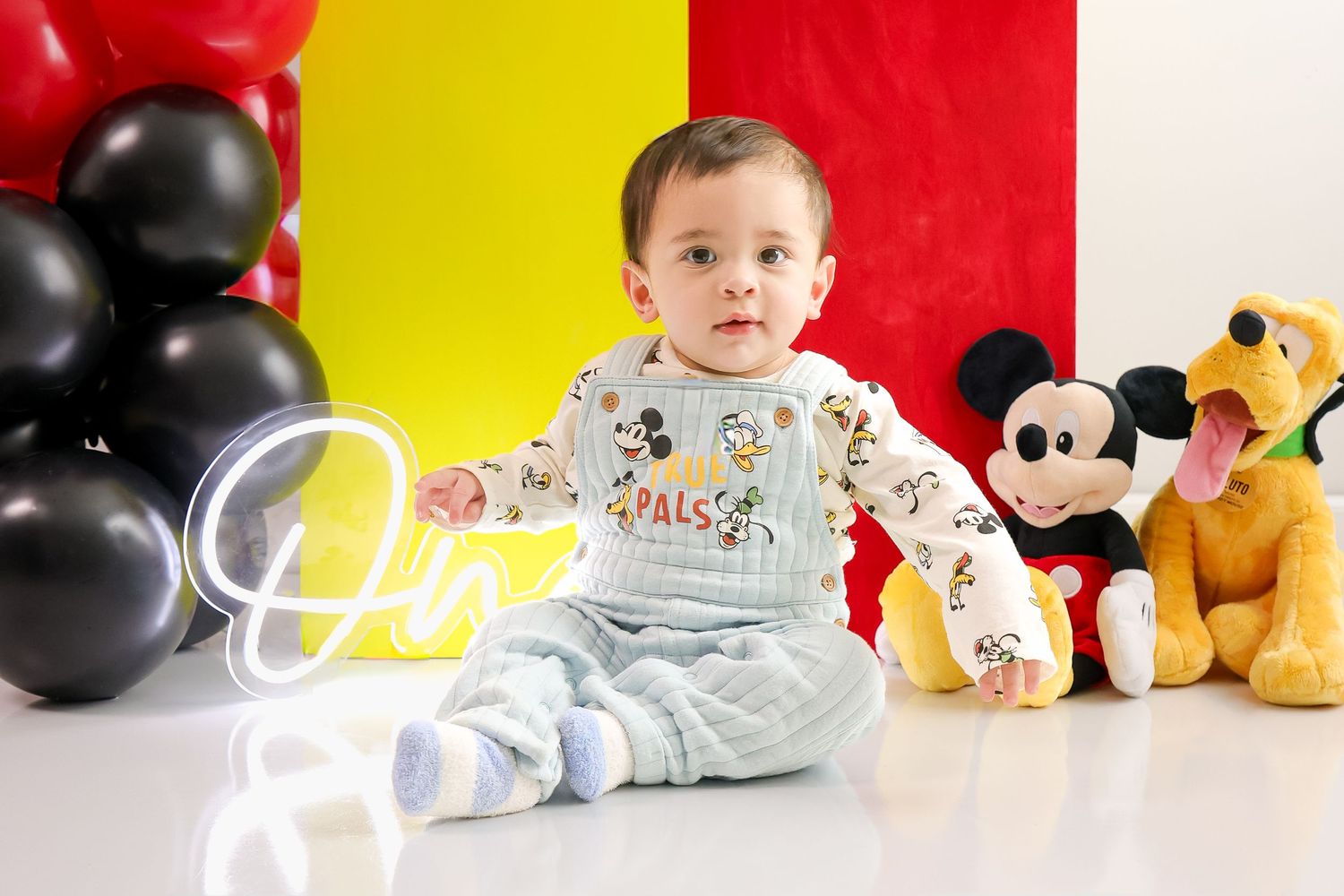 Mickey Mouse themed baby photoshoot setup with red and black balloons, plush toys and white background.
