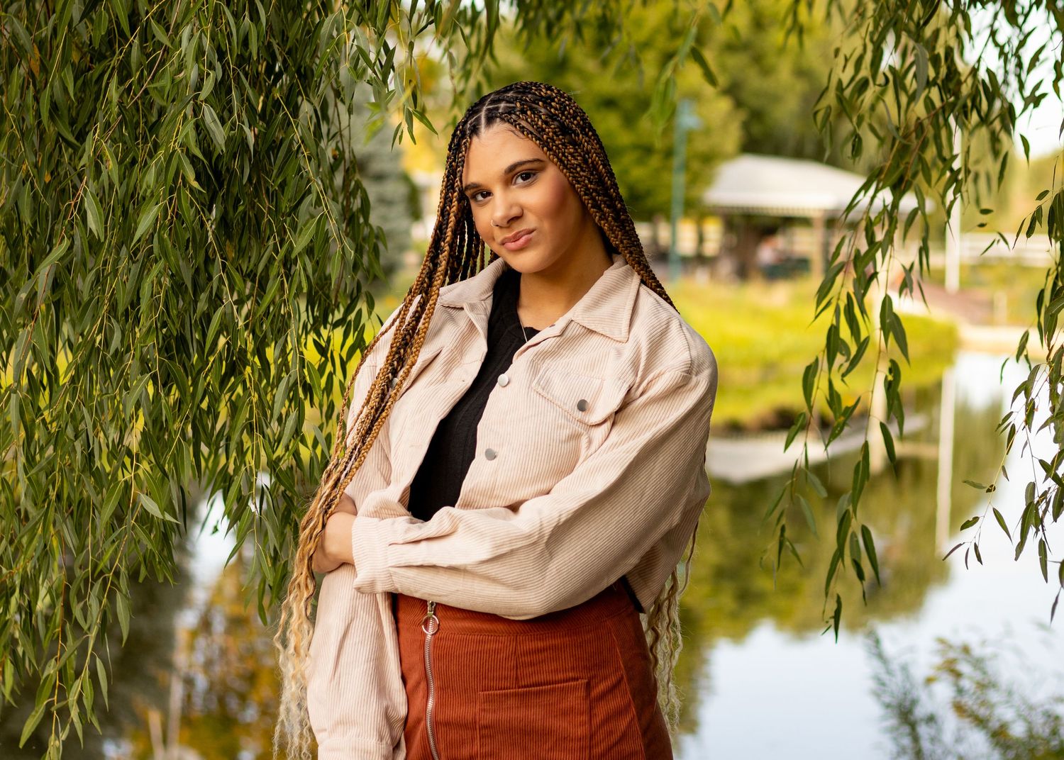 Stylish woman in beige jacket and corduroy skirt posing under willow tree near pond.