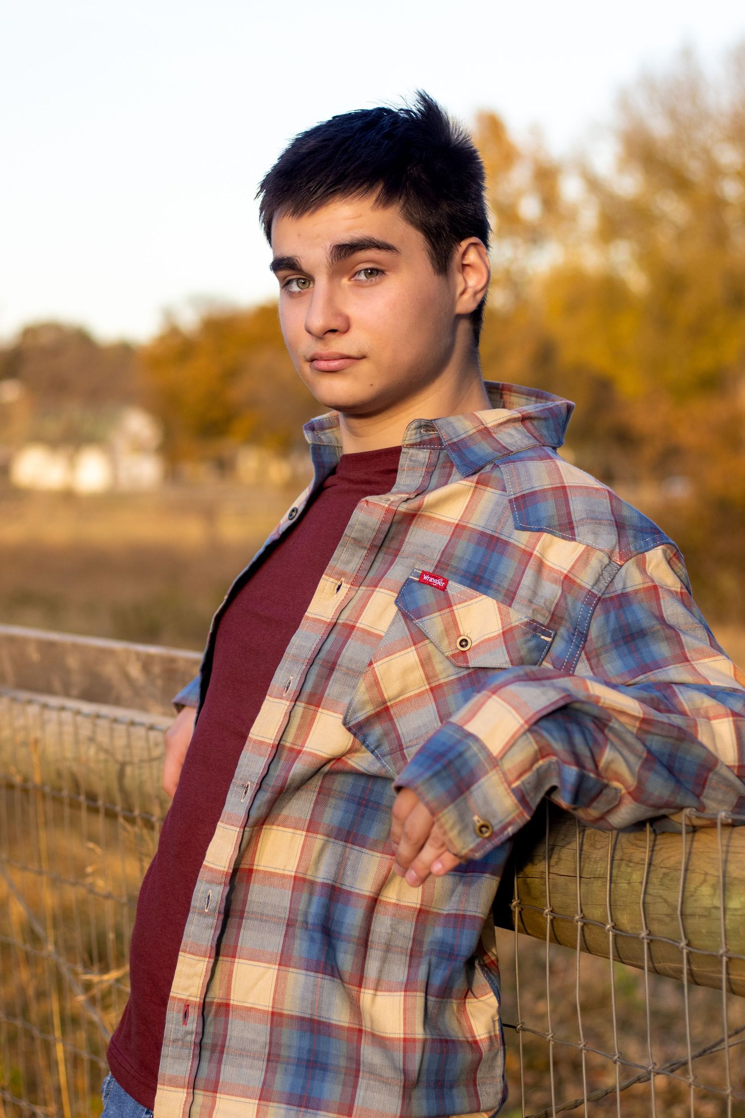 A person in plaid flannel and burgundy shirt leans against a wooden fence at sunset.