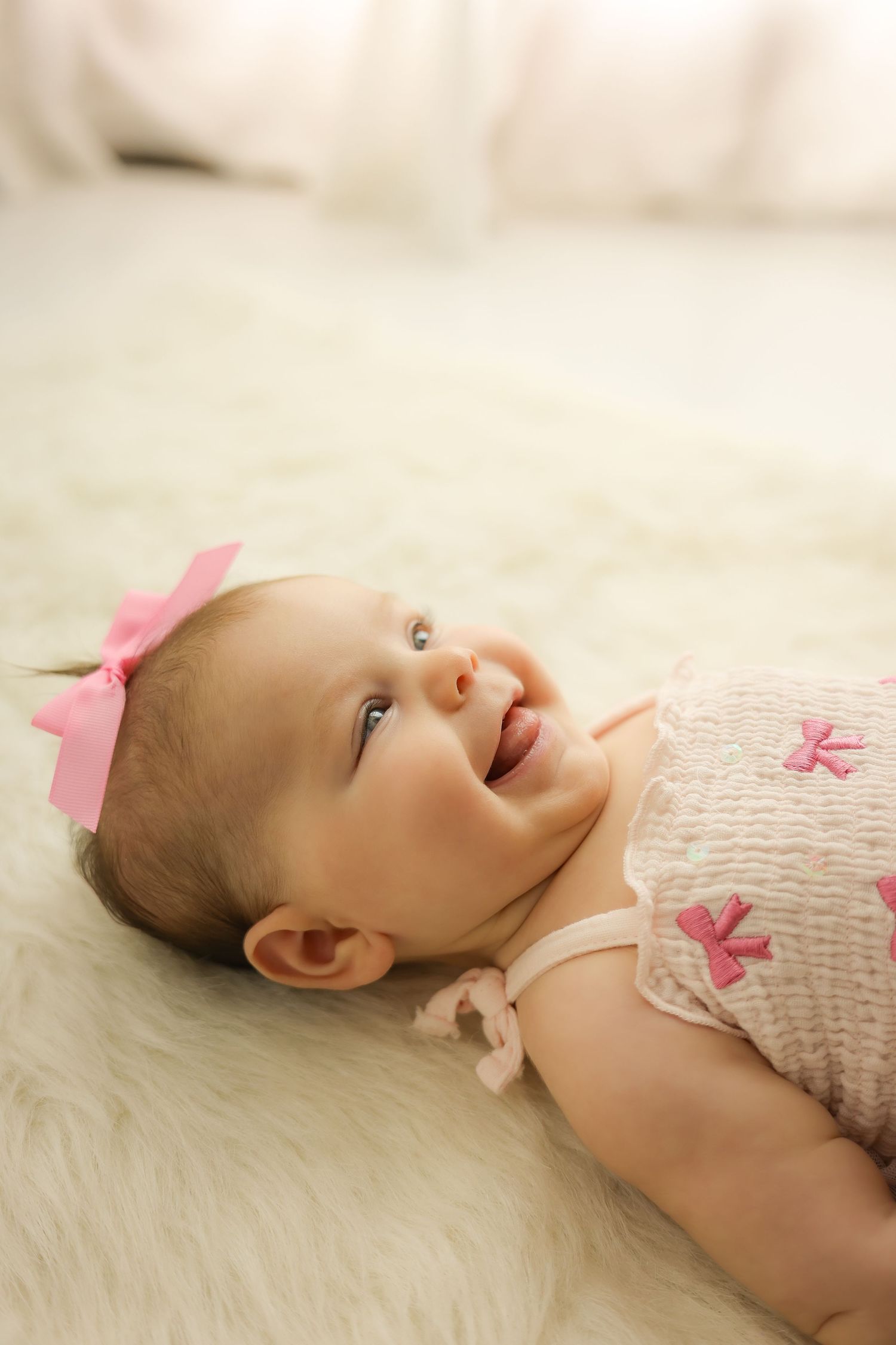 Smiling baby girl wearing a pink bow headband and pink outfit, lying on a fluffy white rug.