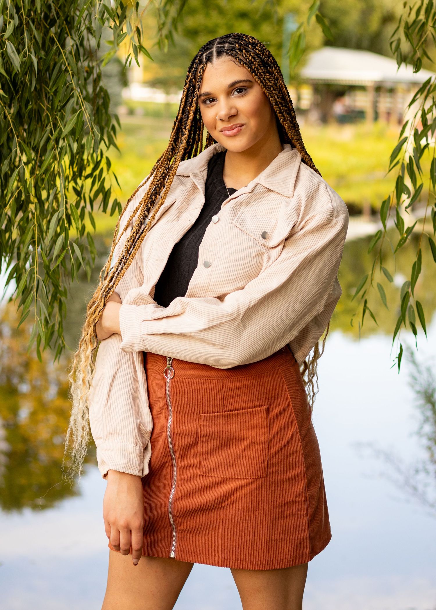 A person in a light denim jacket and rust-colored skirt poses under willow branches near a reflective pond.