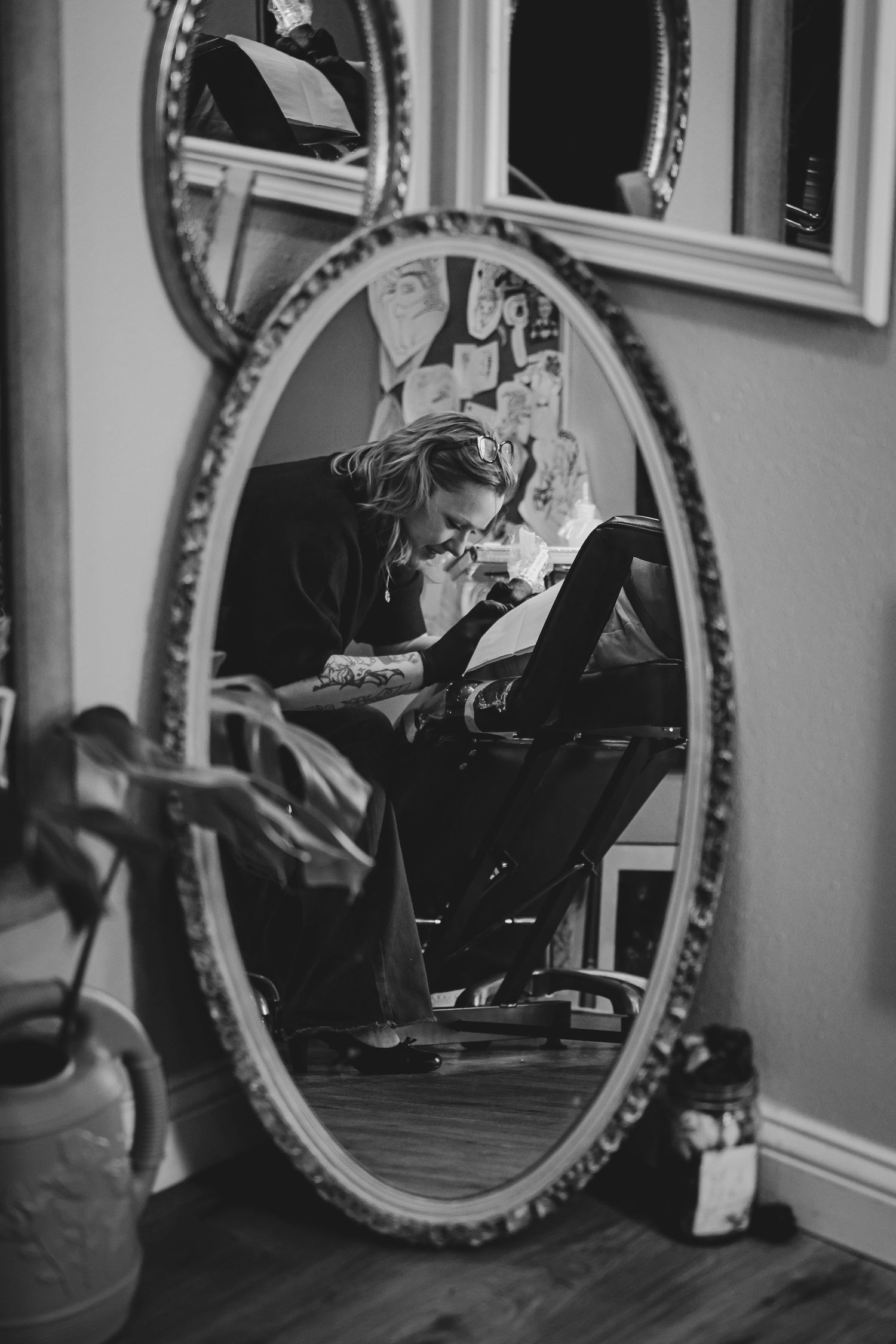 Black and white photo of a tattoo artist working, reflected in an ornate oval mirror in a tattoo studio.