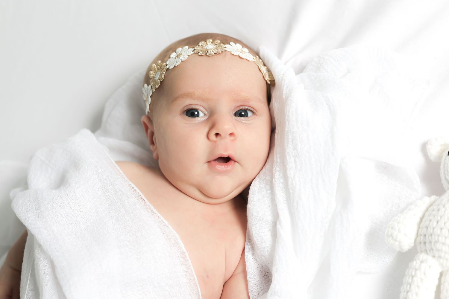 Baby wearing floral crown lying on white bedding with bright expression.