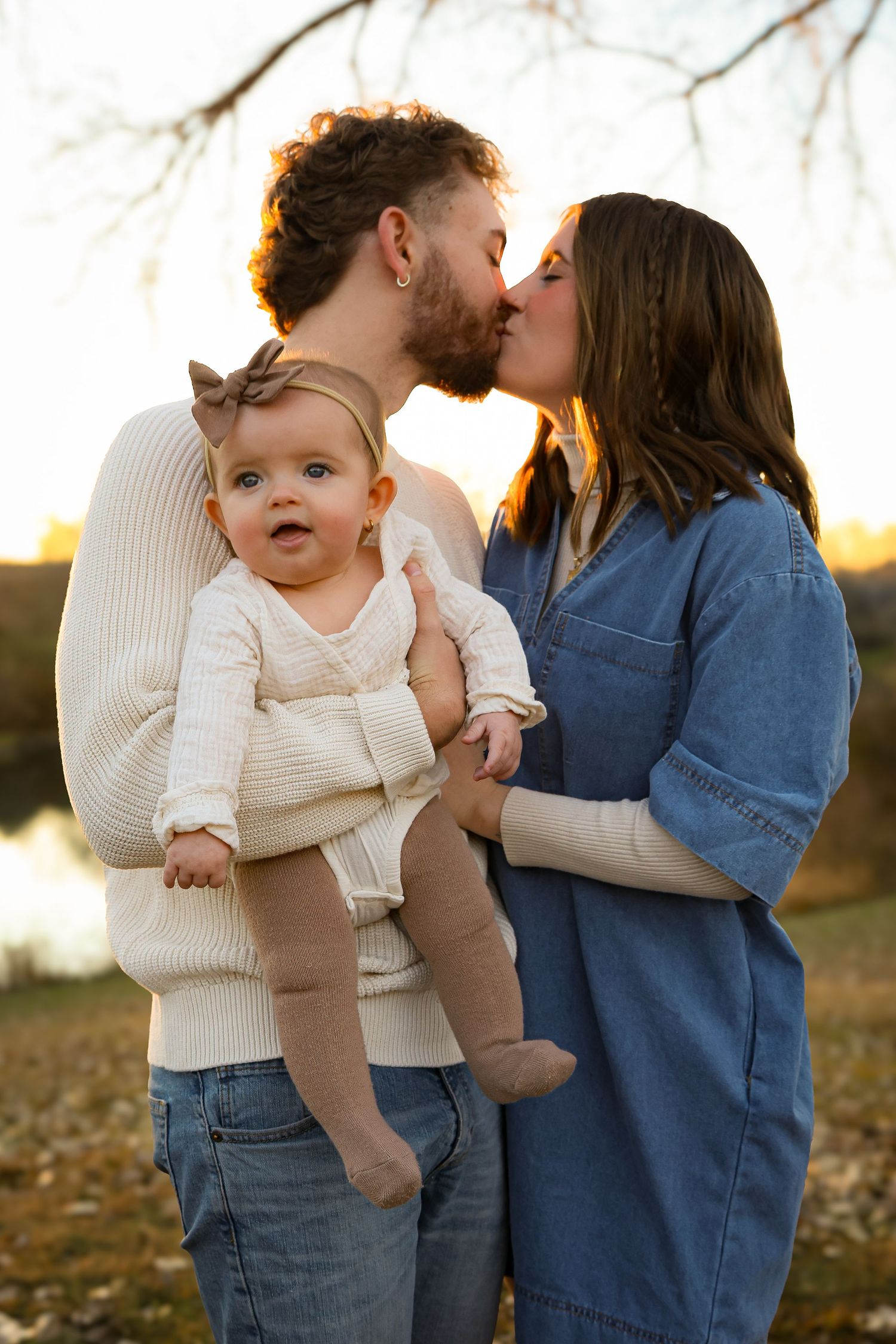 A couple in denim and cream sweaters share tender moments with their baby outdoors during golden hour.