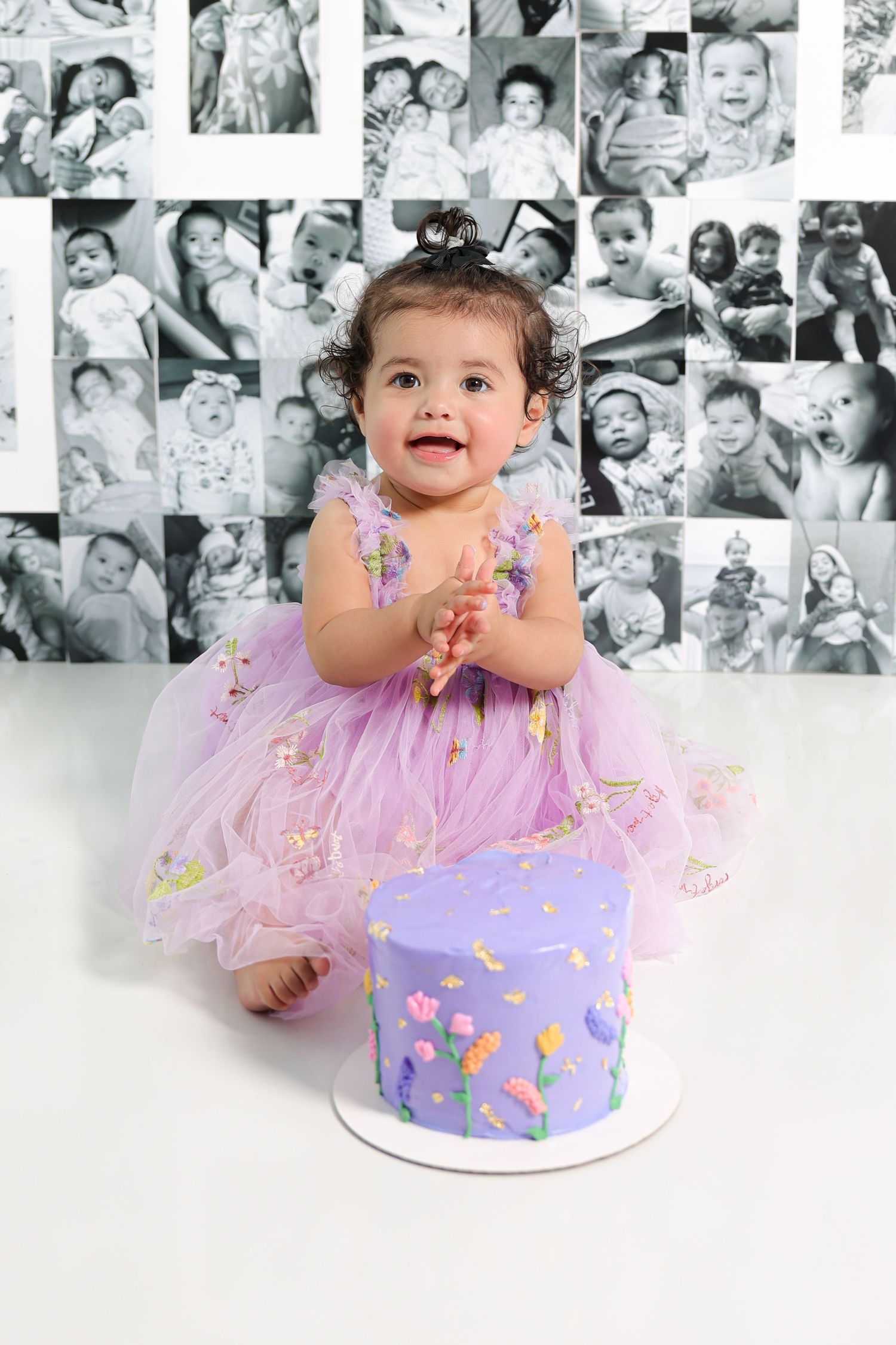 Baby in purple dress reaches for purple birthday cake against black and white photo collage wall.
