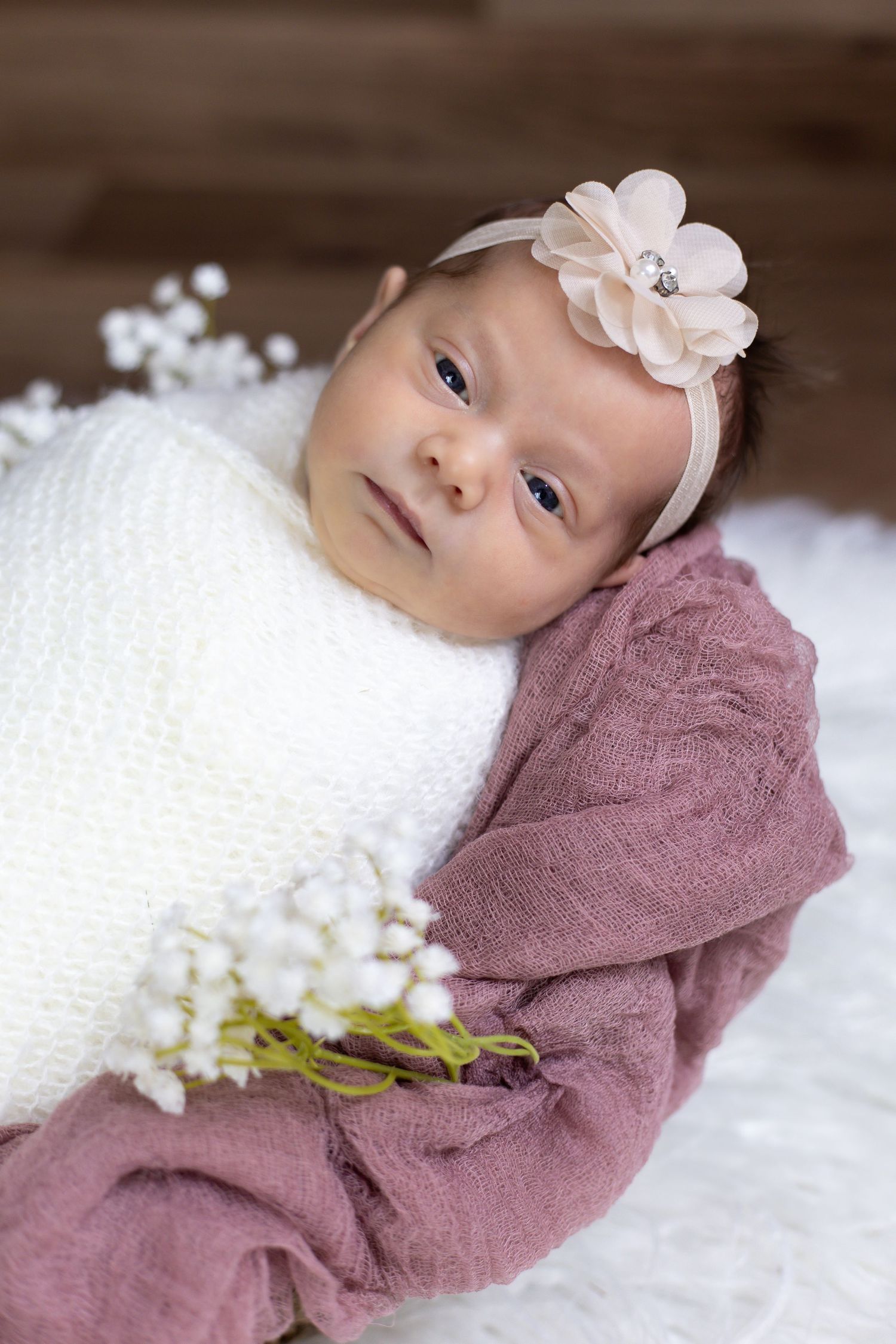 Baby wearing floral headband swaddled in white against mauve fabric with white floral accents.