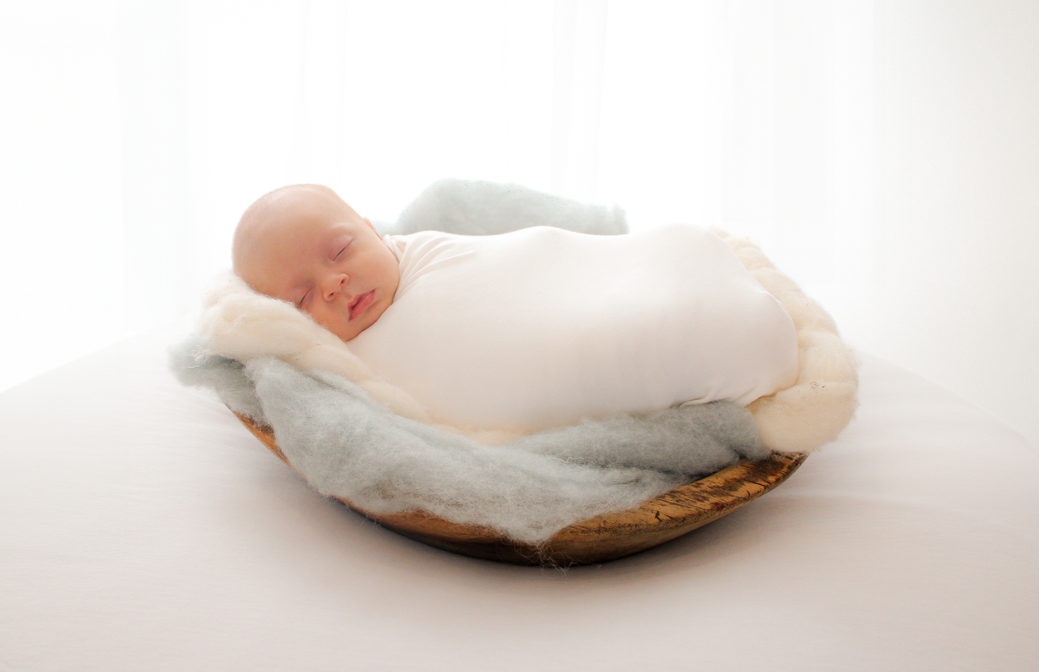 Newborn baby wrapped in white fabric lying in a rustic wooden bowl against white background.