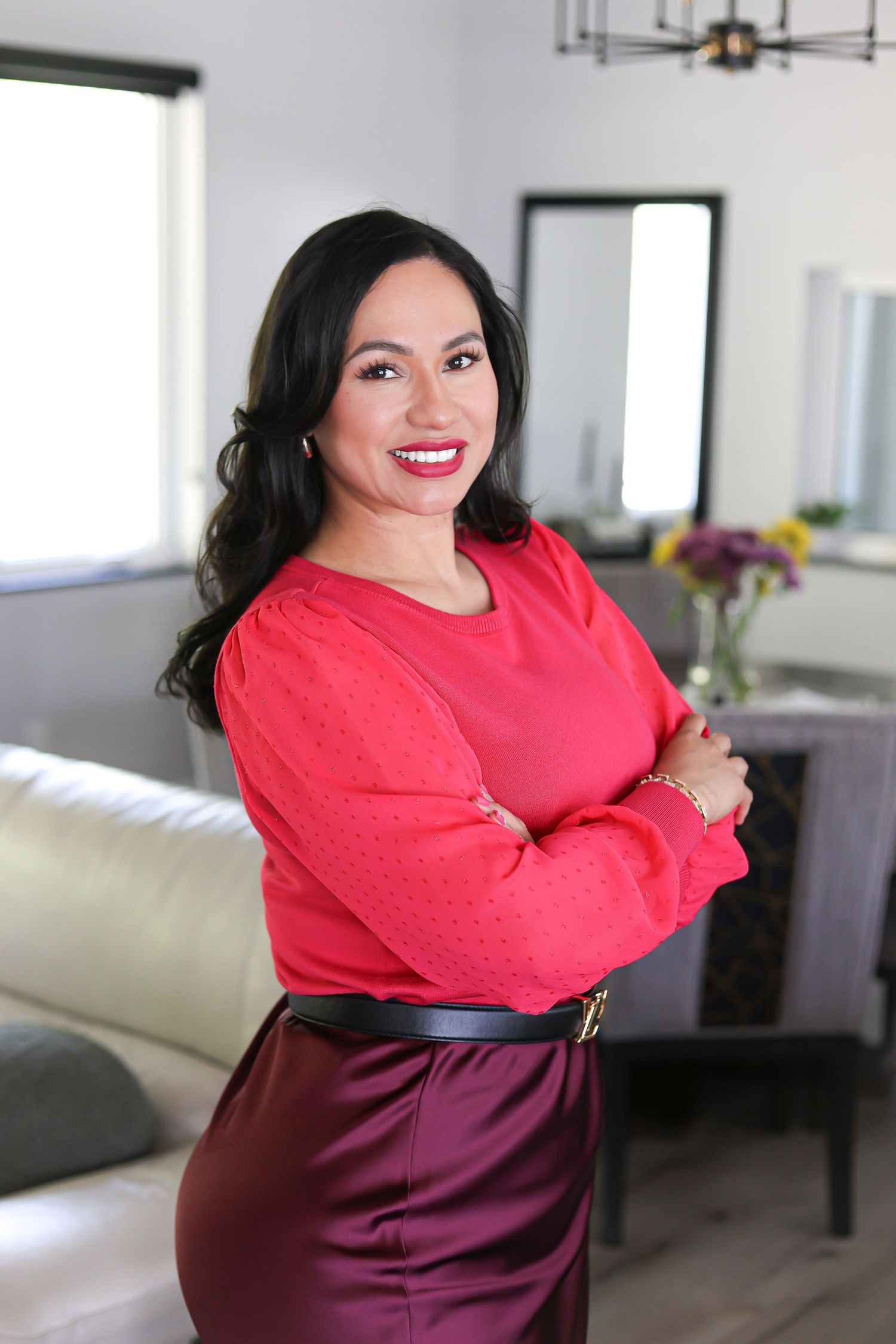 Professional woman in red blouse and burgundy skirt smiling with arms crossed in a modern interior setting.