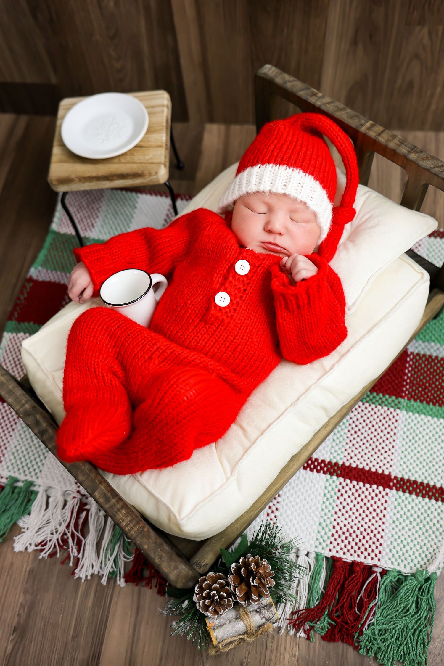 Sleeping infant wearing bright red knitted hat and outfit rests on white pillow with plaid blanket and pinecone decor.
