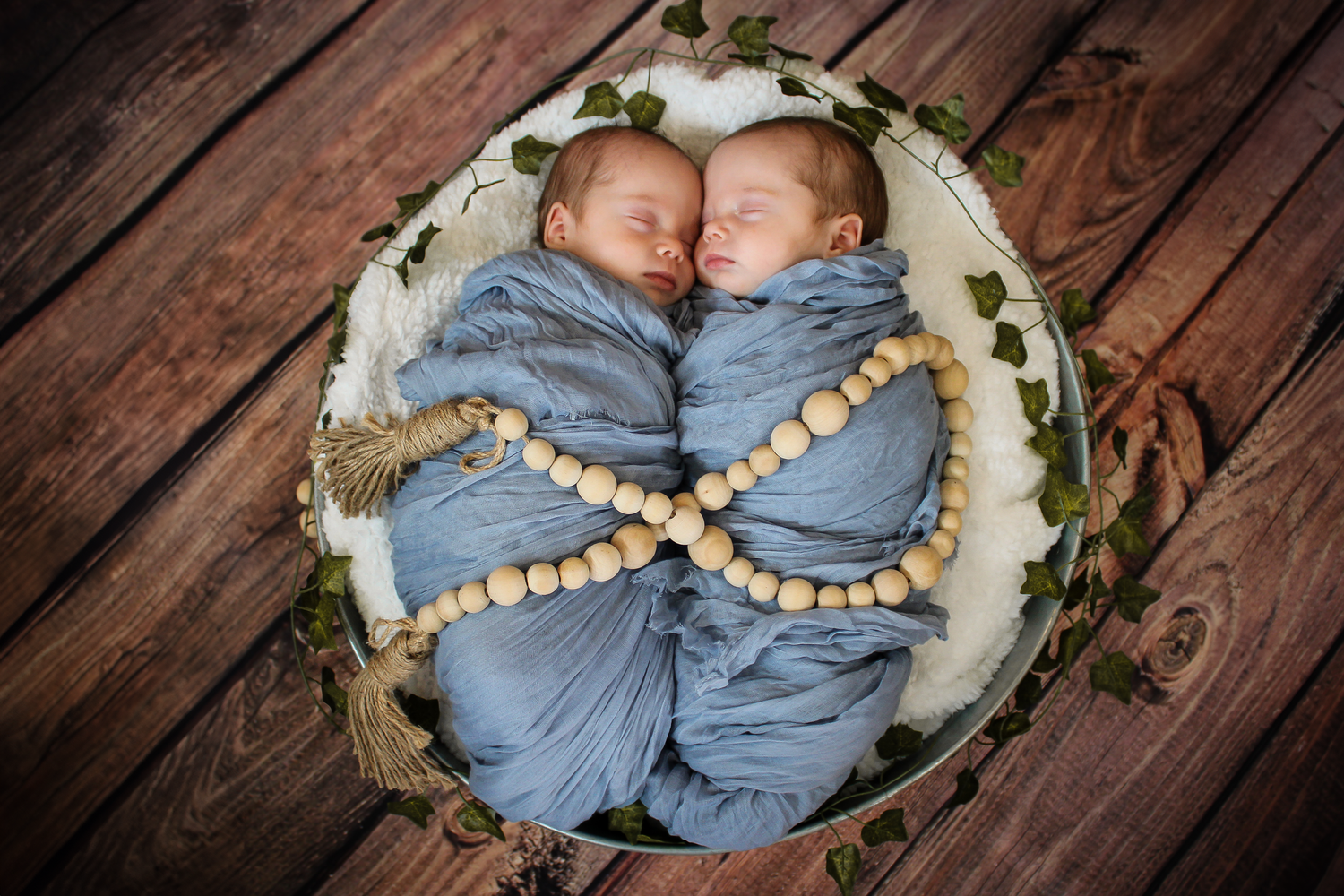 Twin babies swaddled in blue blankets lying in circular basket with floral decor.