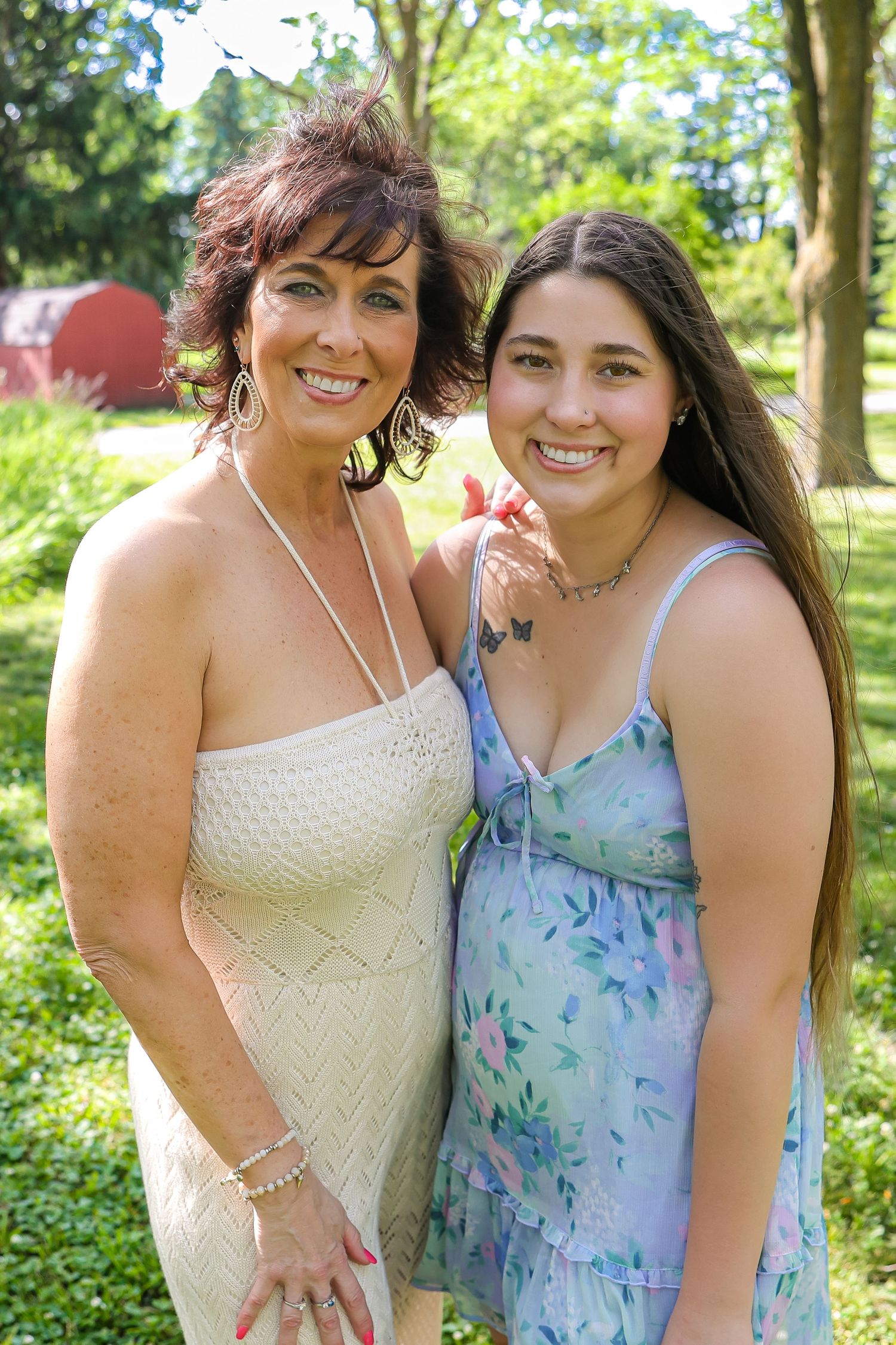 Two people in summer dresses pose together in a lush green outdoor setting.
