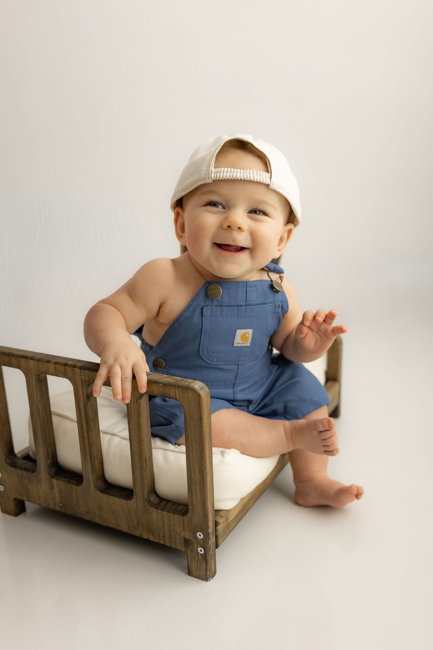 Smiling baby in blue Carhartt overalls and white cap sits on a miniature wooden bed during a photo session.
