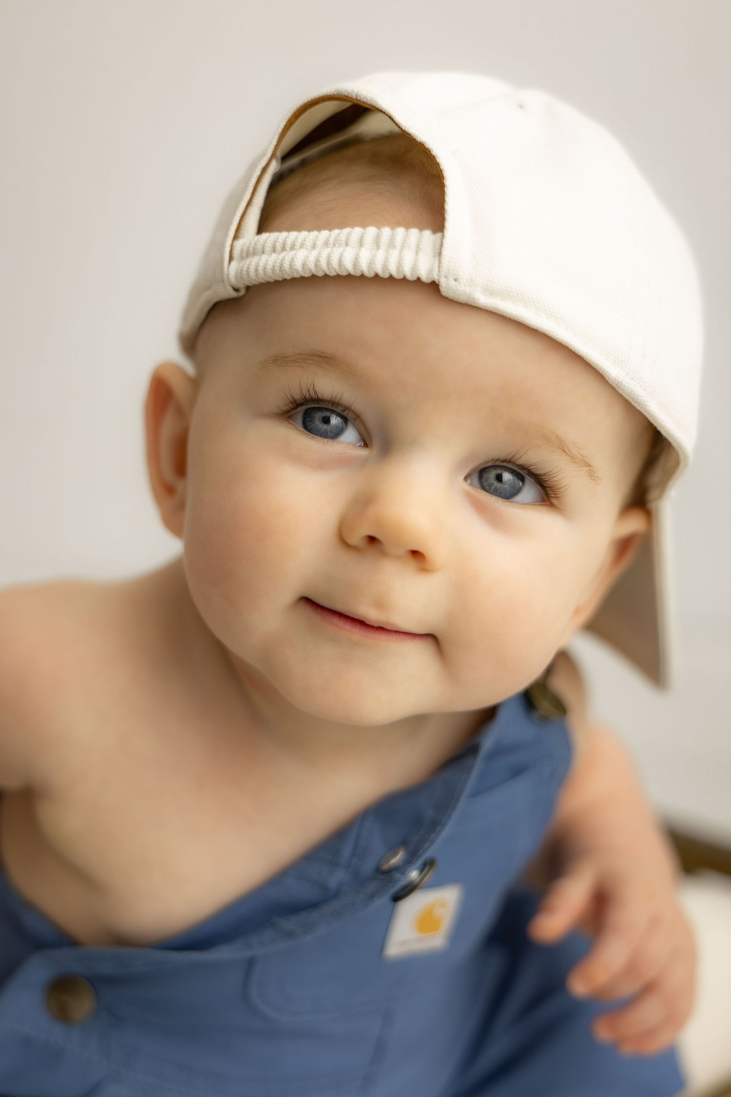Adorable baby with blue eyes wearing a white backwards cap and blue Carhartt overalls, smiling softly.