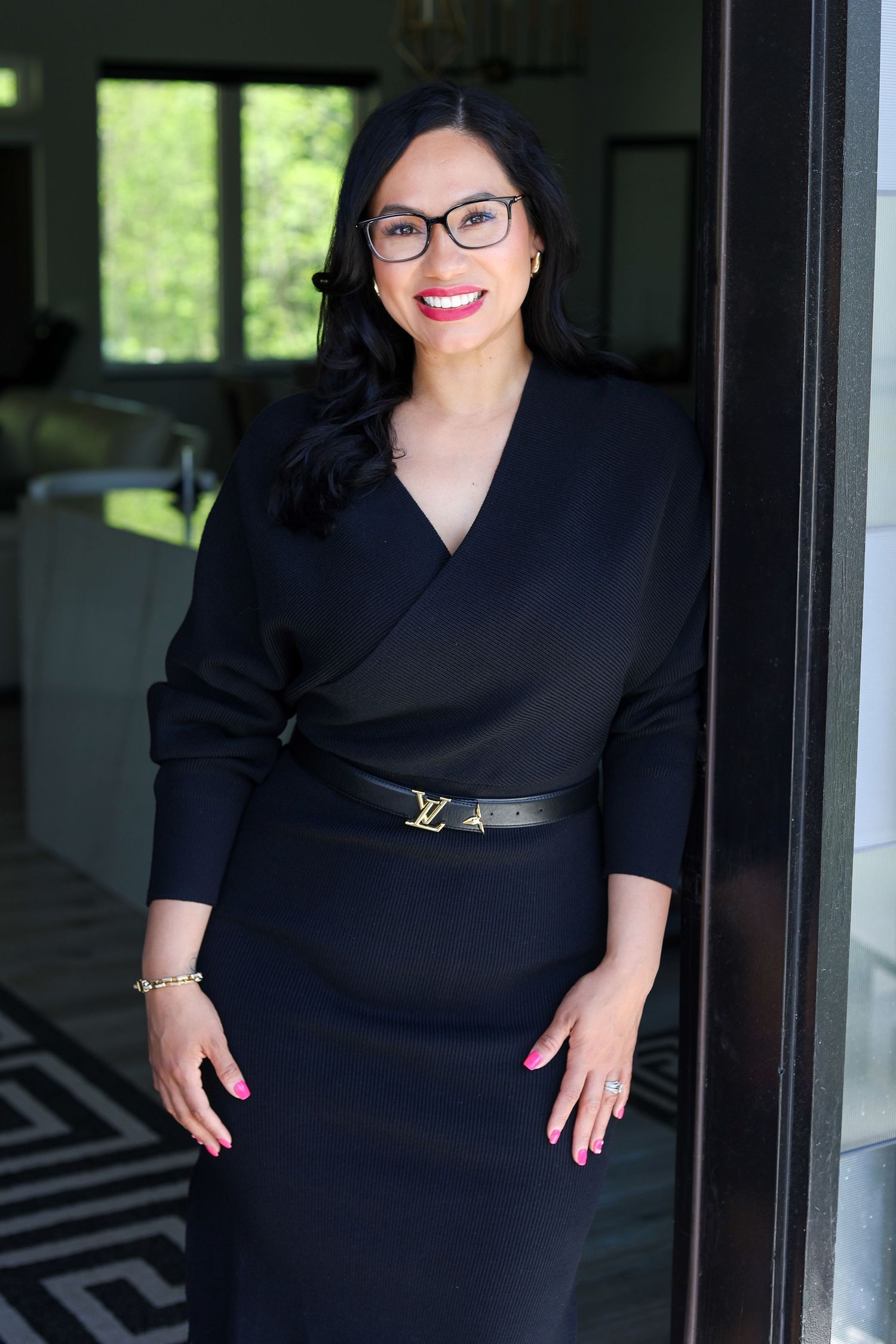 Woman in black wrap dress with Louis Vuitton belt and glasses smiling confidently in an elegant interior doorway.