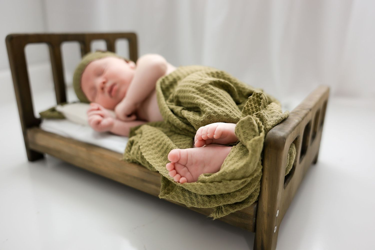 A sleeping newborn baby lies peacefully on a wooden bed wearing a cozy knit blanket in warm natural light.