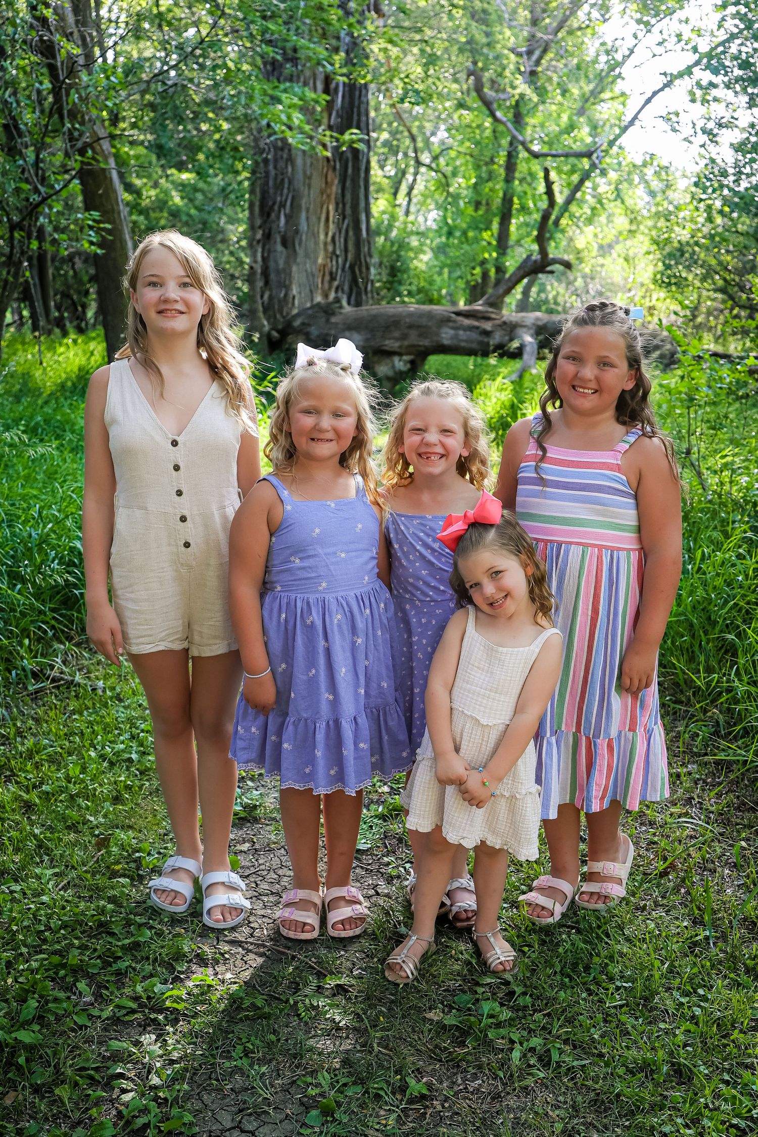Children wearing coordinated summer outfits pose together on a wooded path in natural lighting.