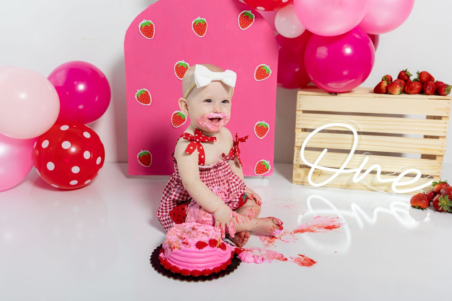 Pink themed cake smash photoshoot setup with strawberry decorations and balloons against a pink backdrop.