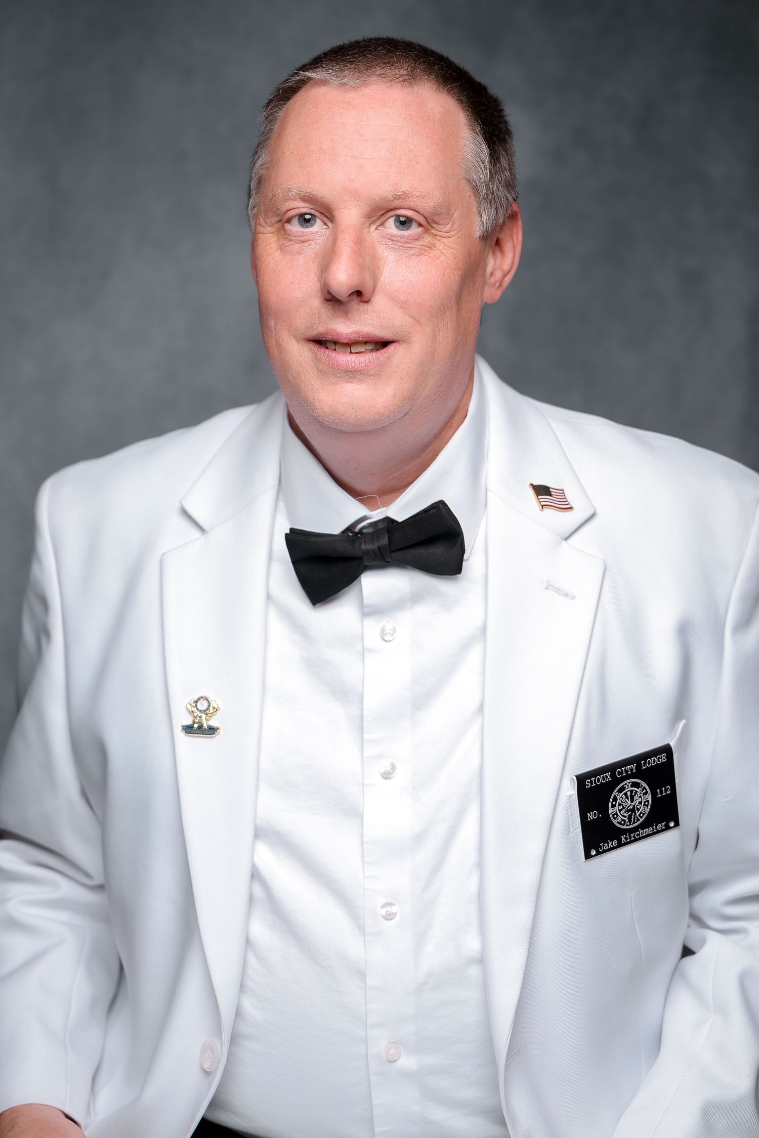 Man in white tuxedo jacket with black bow tie and American flag pin poses for formal portrait photo.