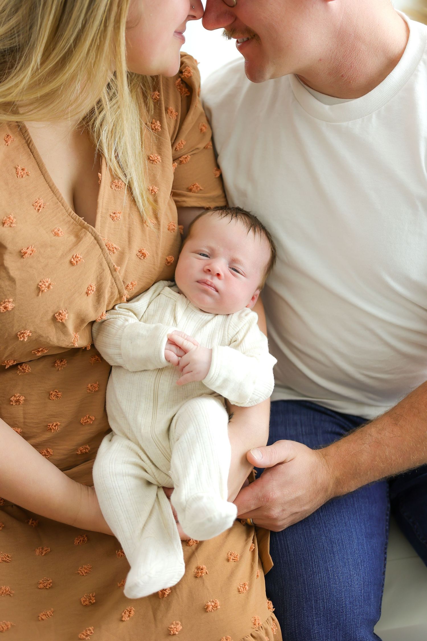 A sleeping newborn baby in white clothing rests between two adults in casual attire.