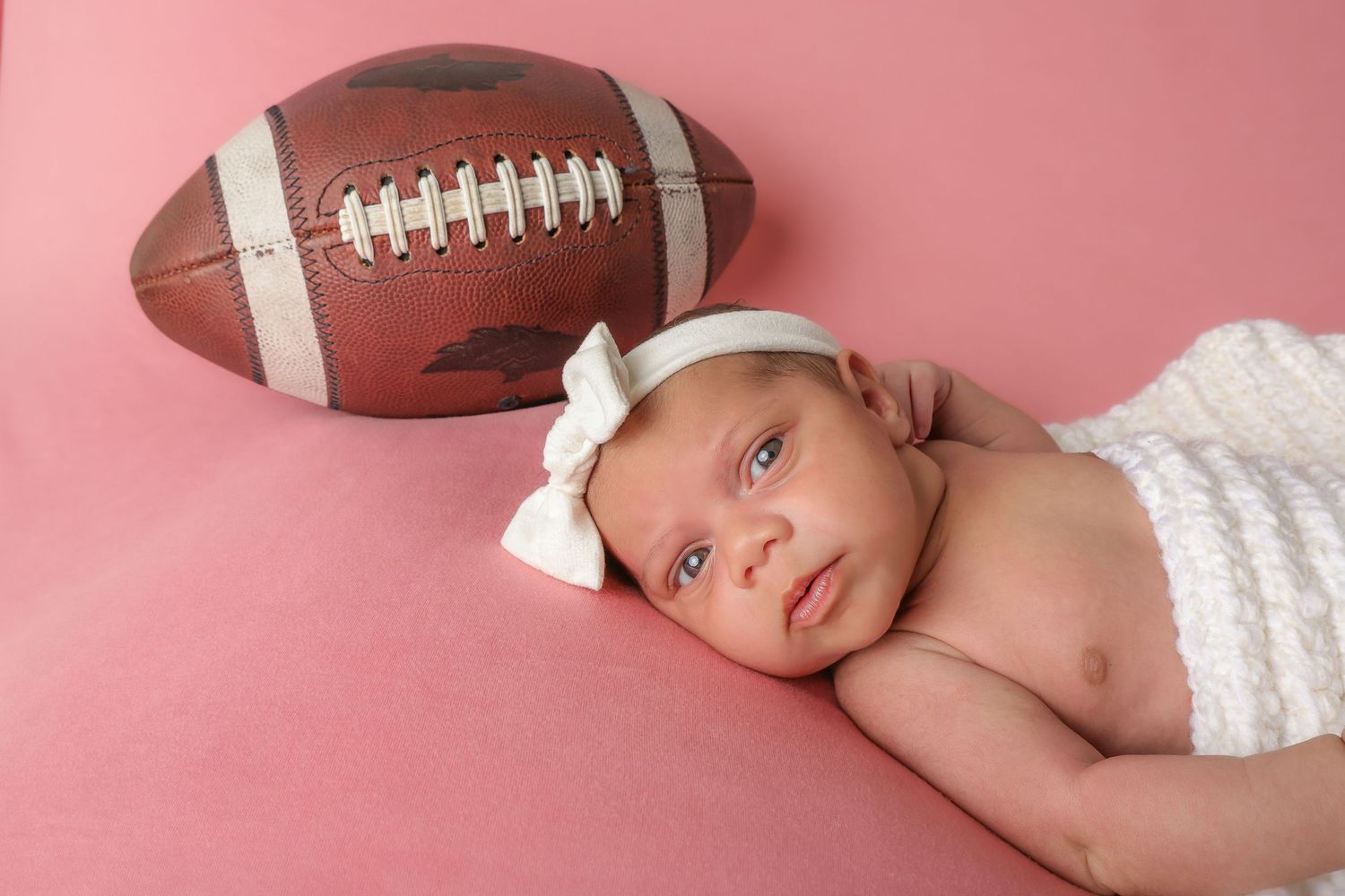 A newborn baby lies on a pink backdrop with a football prop in a professional portrait setting.