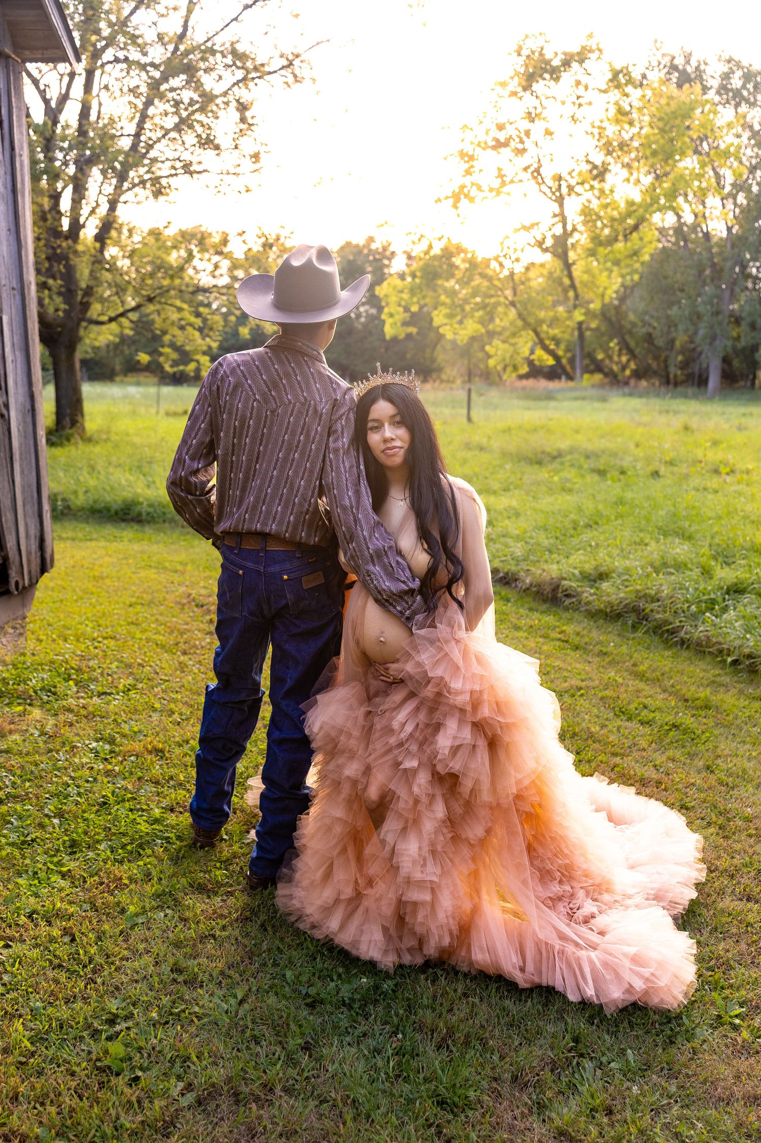 A couple poses in a pastoral setting at sunset, with a flowing peach-colored gown and western attire creating a romantic scene.
