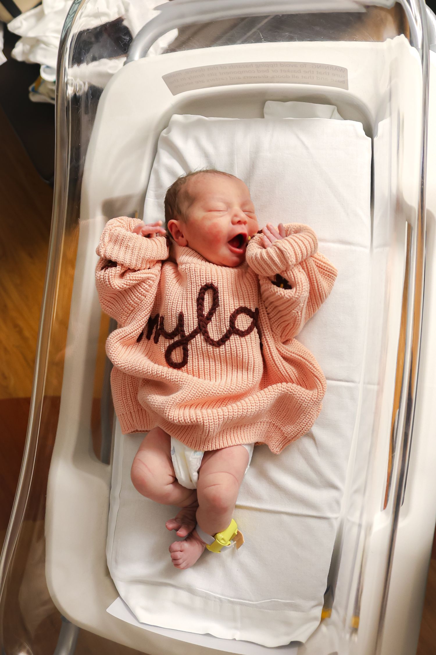 Overhead view of newborn wearing peach sweater in clear hospital bassinet.