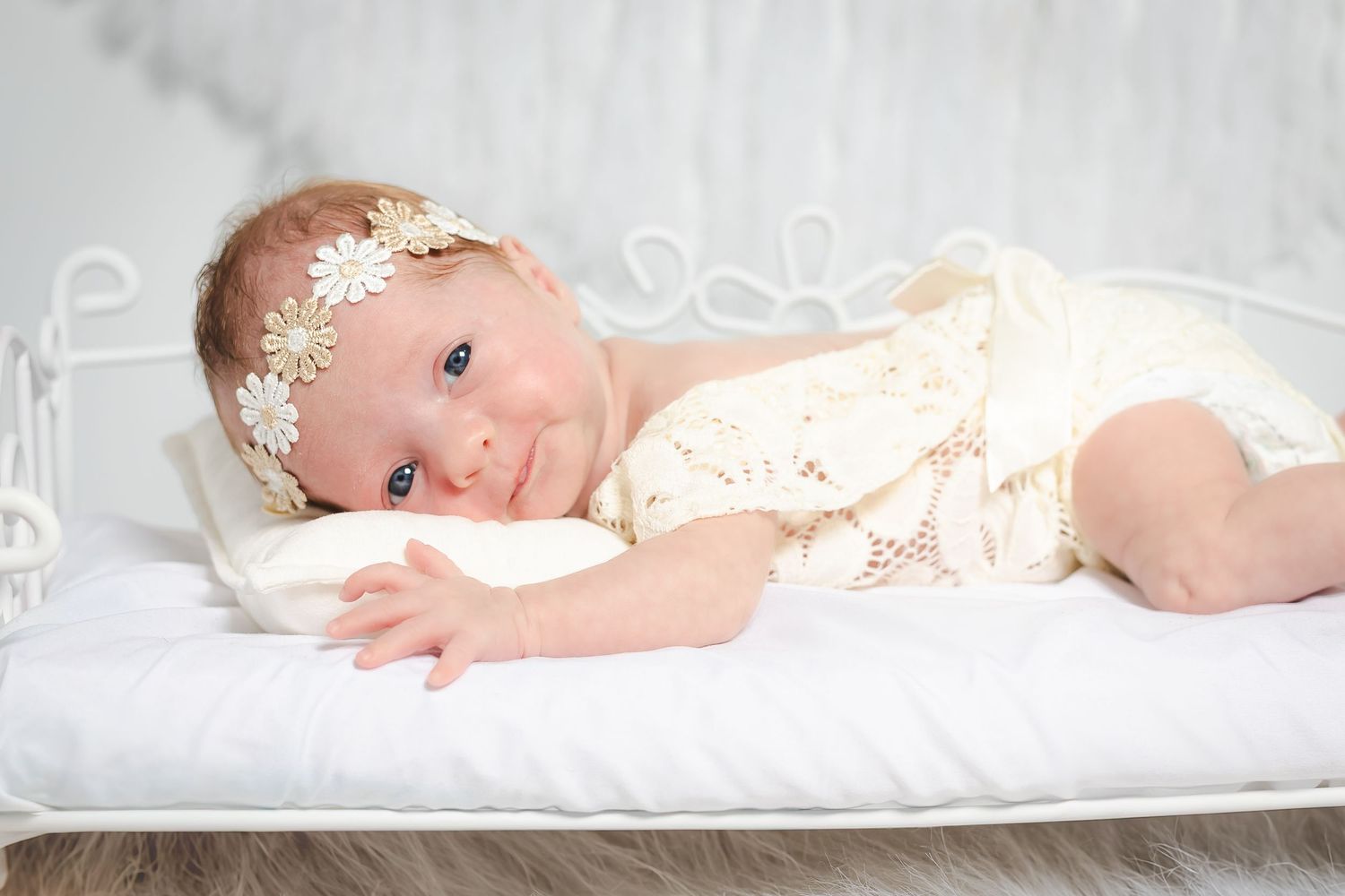 Baby in white lace dress with delicate headband lying on soft white bedding.