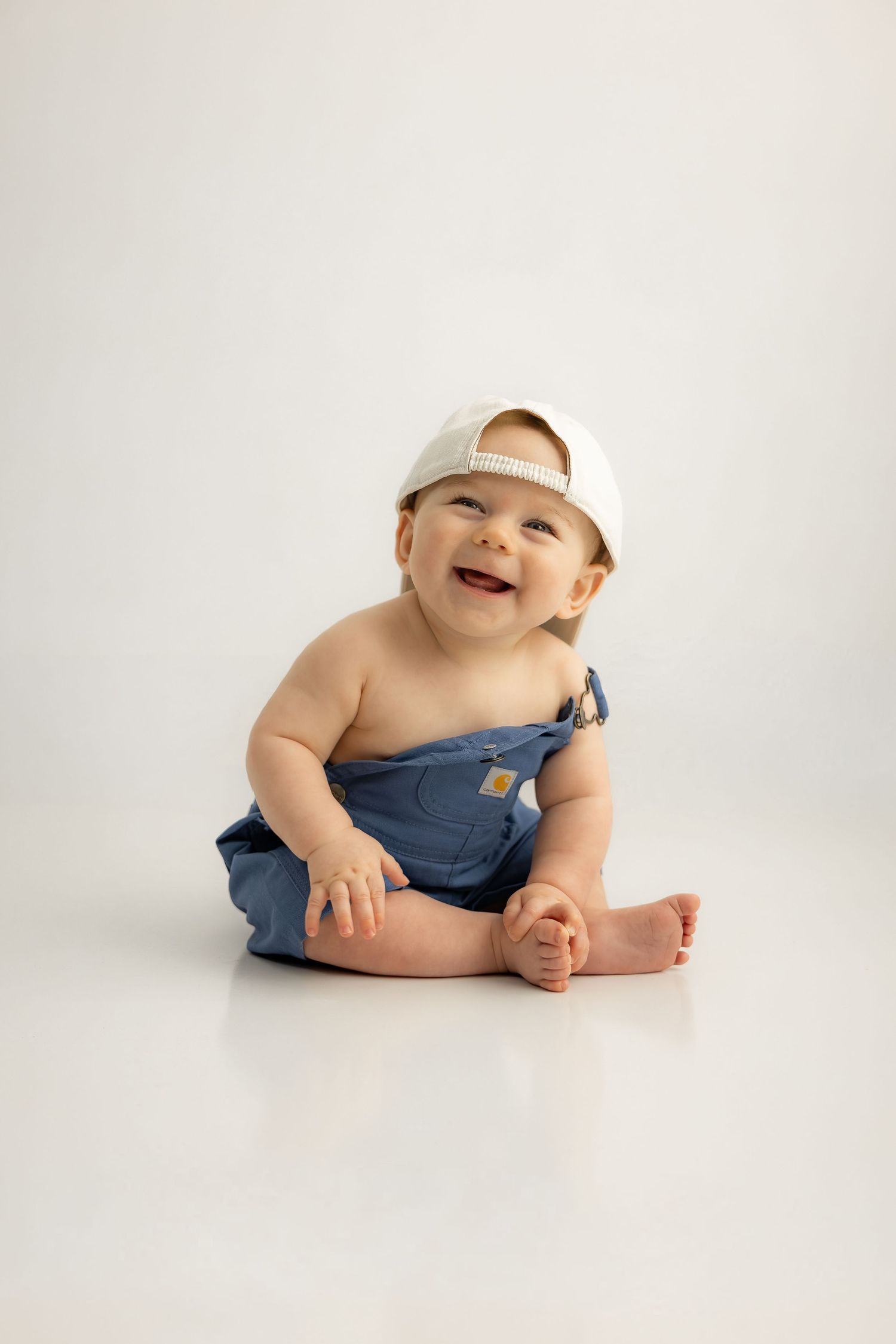 Smiling baby in blue denim overalls and white baseball cap sitting on white background.