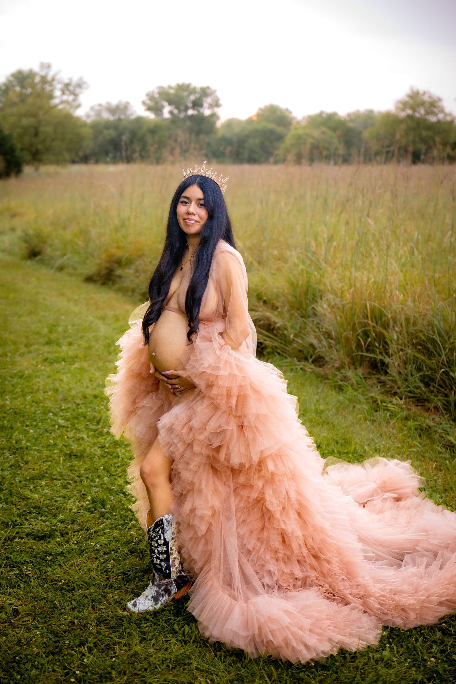 A flowing peach tulle gown creates a dreamy silhouette against a green meadow landscape in an outdoor photo series.