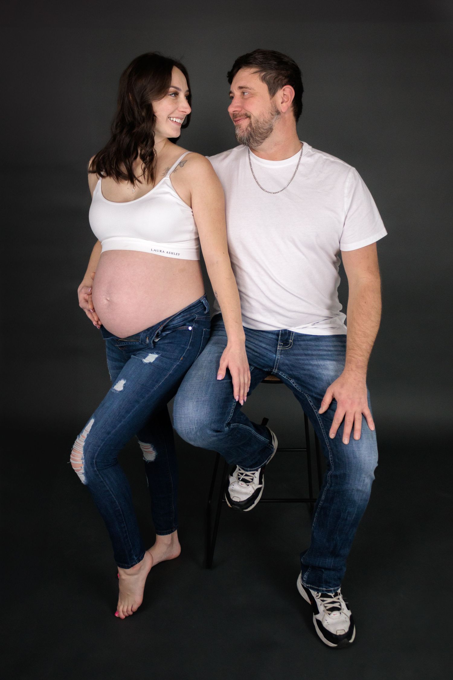 Casual maternity photoshoot with couple in white shirts and jeans against dark backdrop.