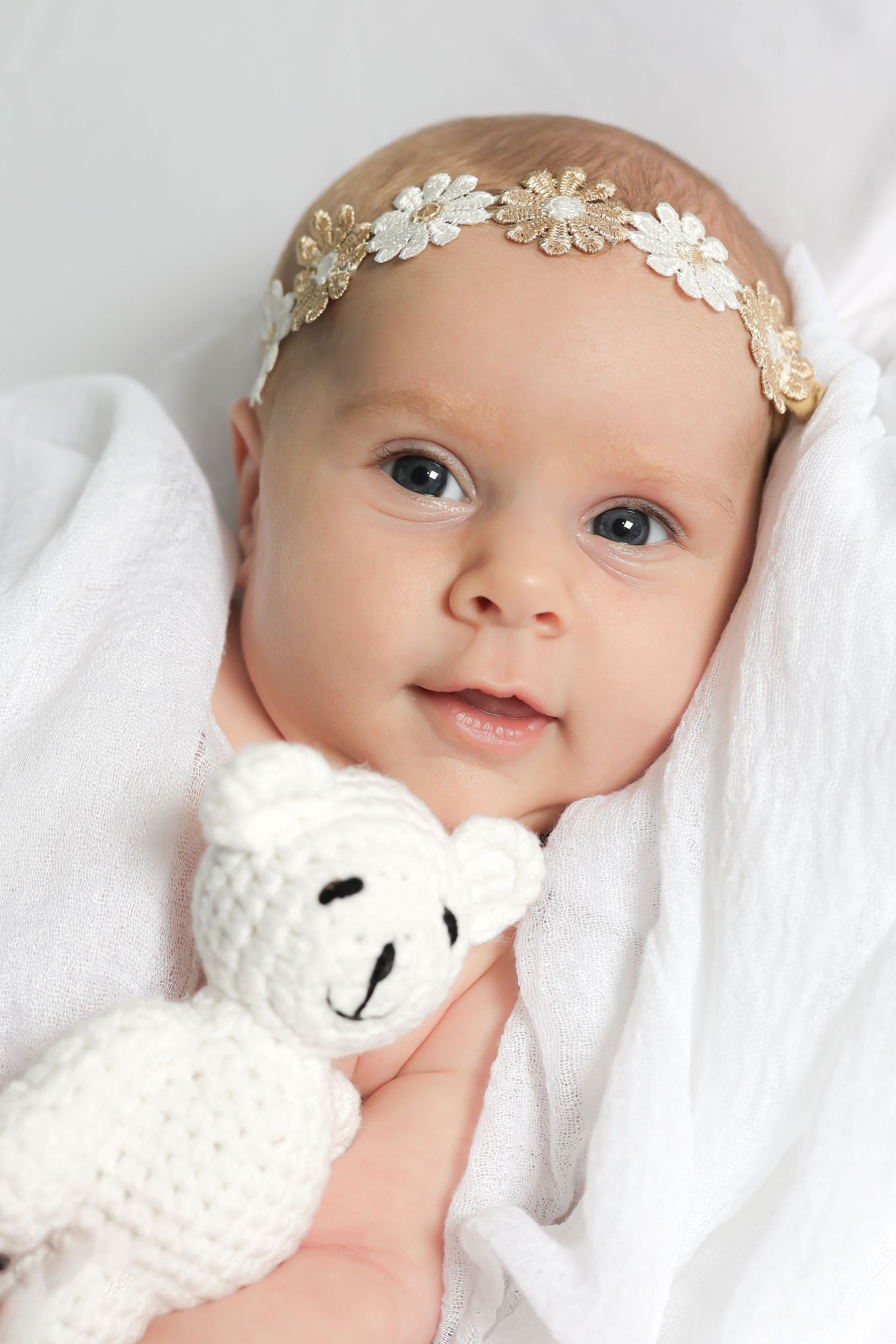 Newborn wearing a delicate floral headband and white knit outfit lying on white bedding.