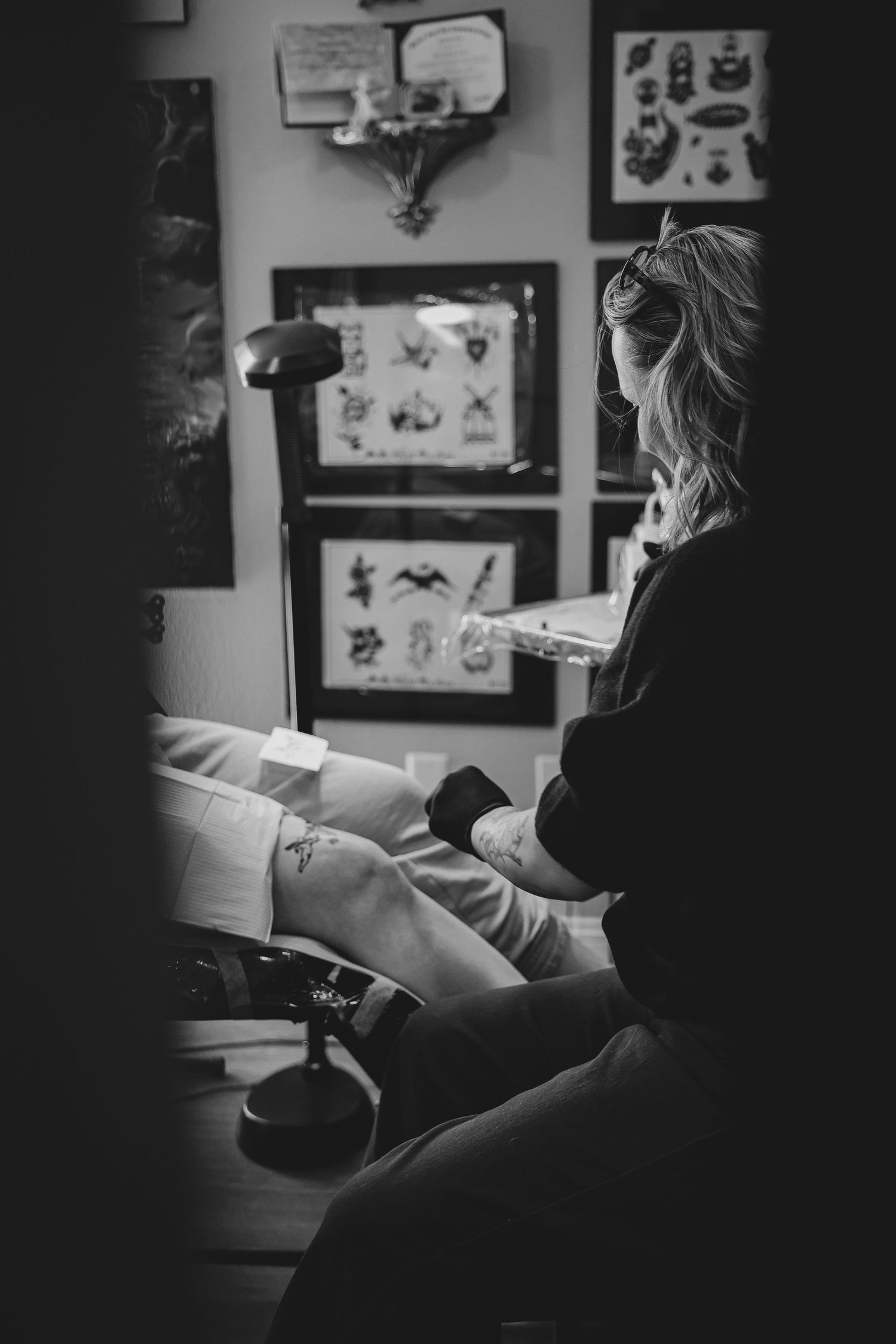 Black and white photo of a female tattoo artist working on a client's leg in a studio with flash art on the walls.