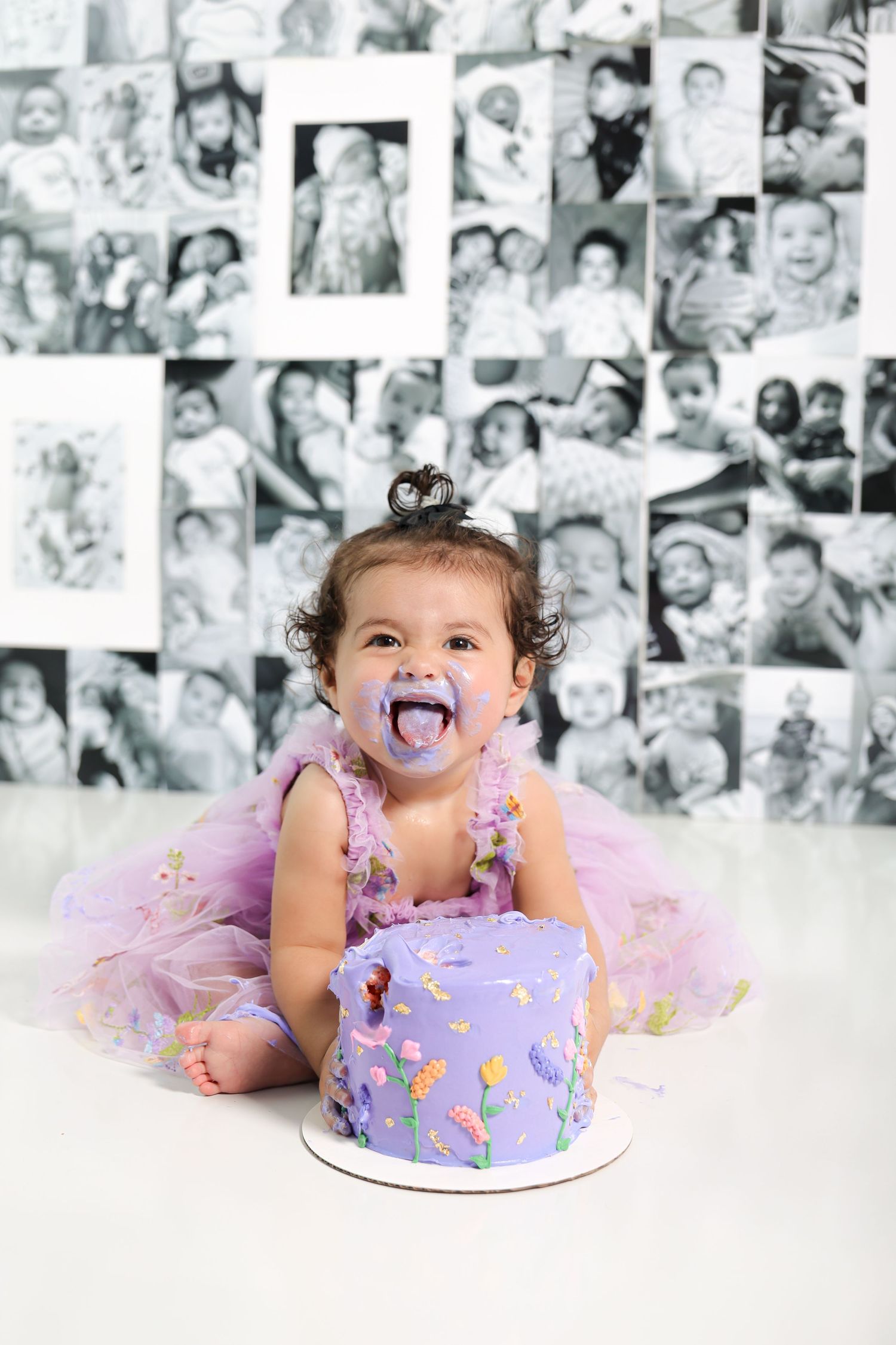 A baby in a purple tutu dress sits with a matching purple birthday cake against a wall of black and white photos.