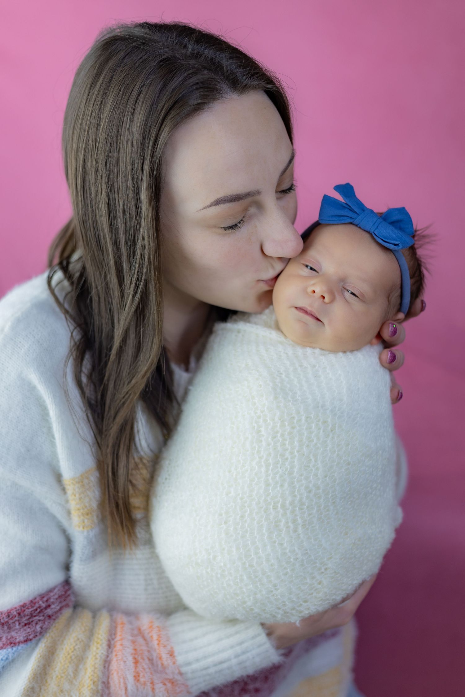 Tender moment captured against pink background with baby wrapped in white knit blanket.