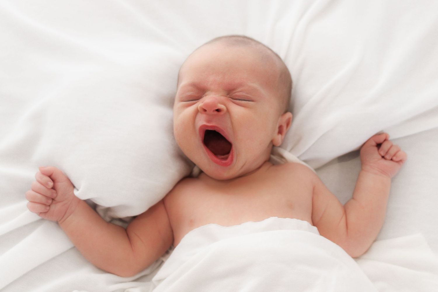 A sleeping newborn baby yawning while lying on white bedding.
