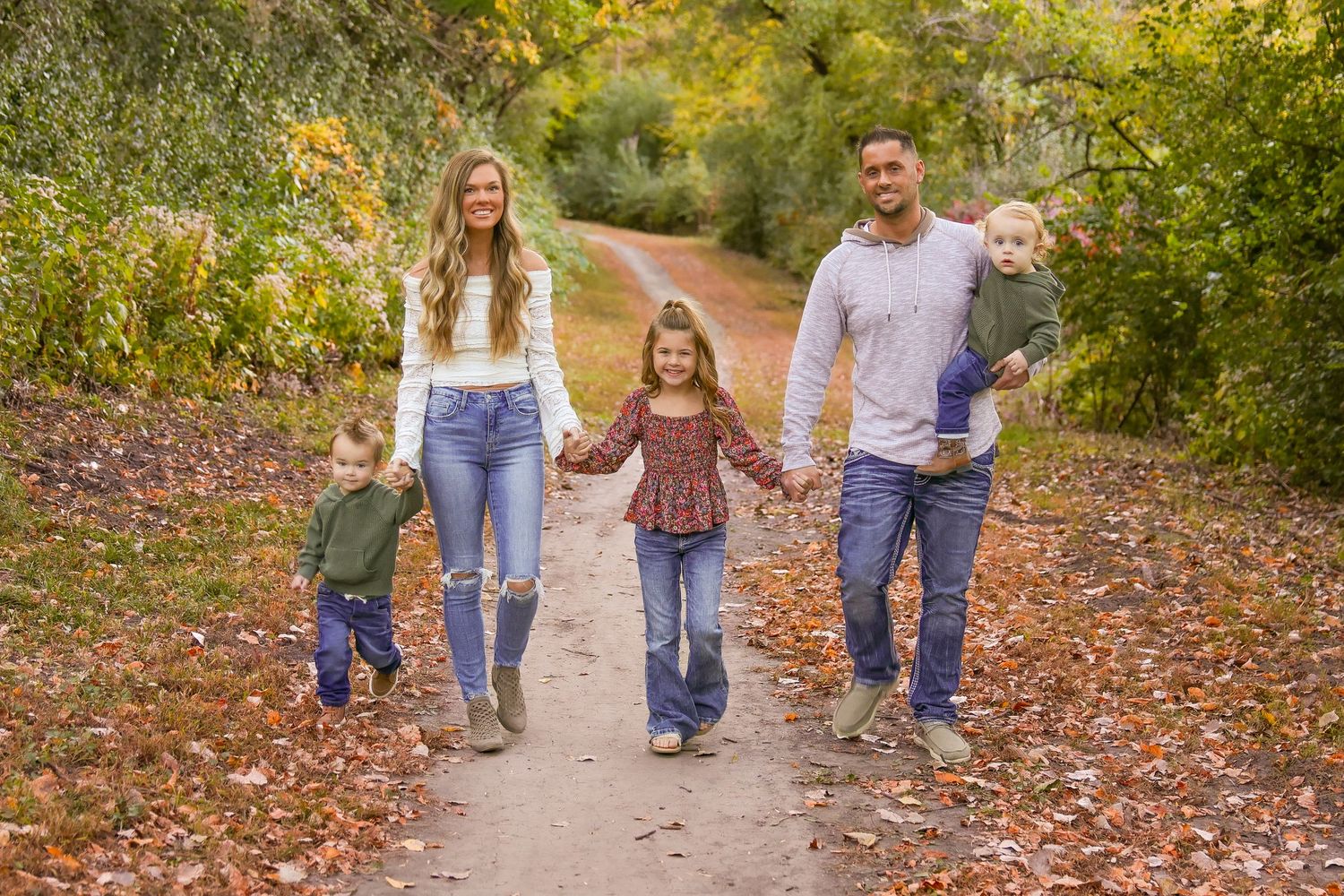 A family walks together on a leaf-covered autumn trail surrounded by golden foliage.