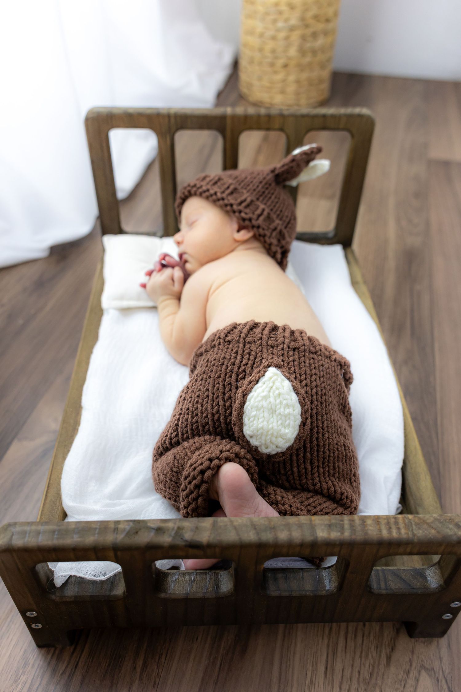 Newborn baby wearing brown knitted bunny outfit lying on wooden bench with white cushioning.