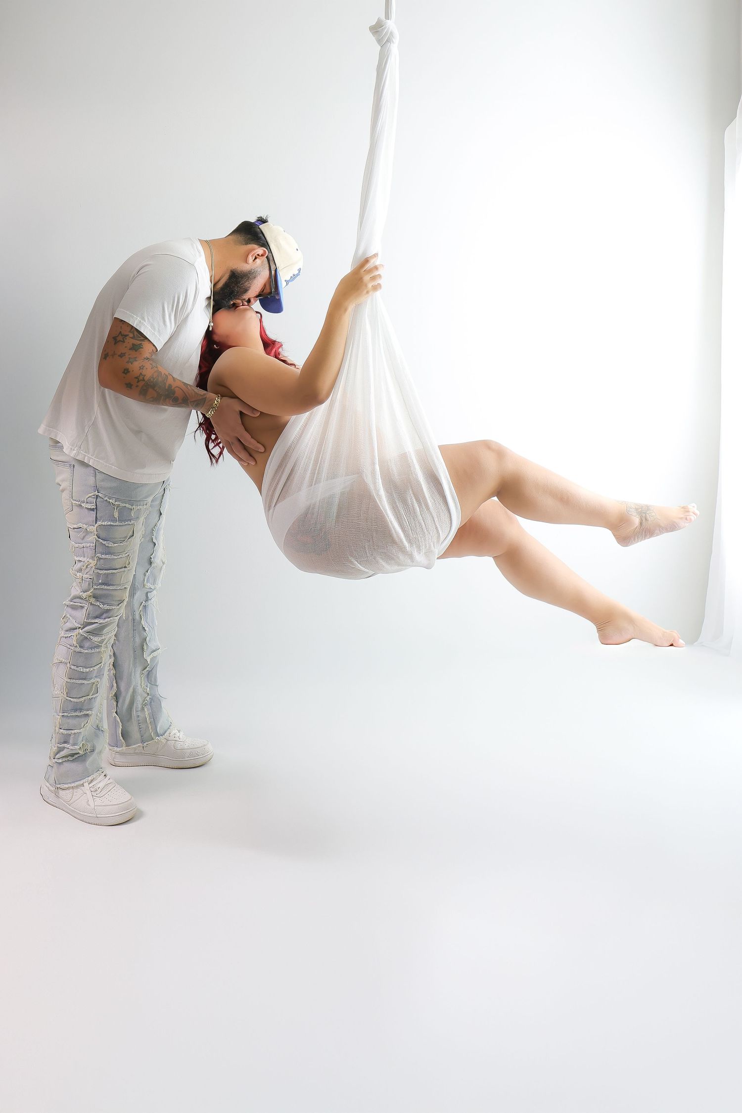 Artistic photo of two people in white, with one suspended in flowing fabric against a bright minimalist background.