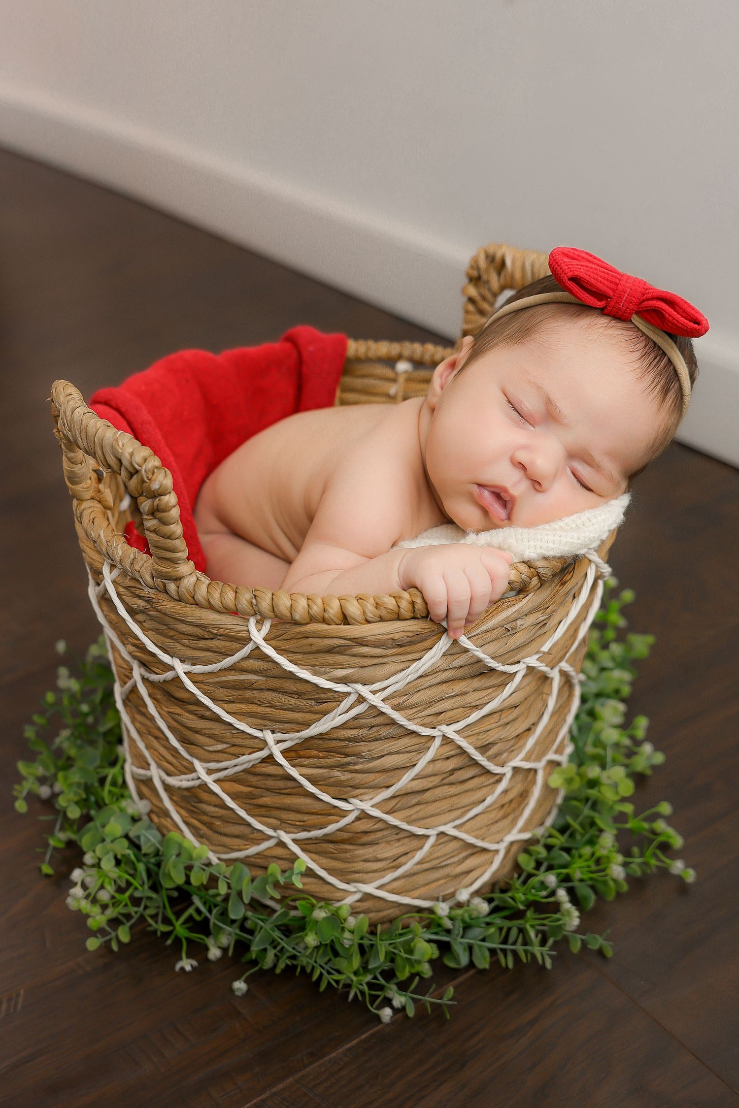 A sleeping newborn nestled in a woven basket decorated with a red bow and surrounded by green foliage on a dark wooden floor.