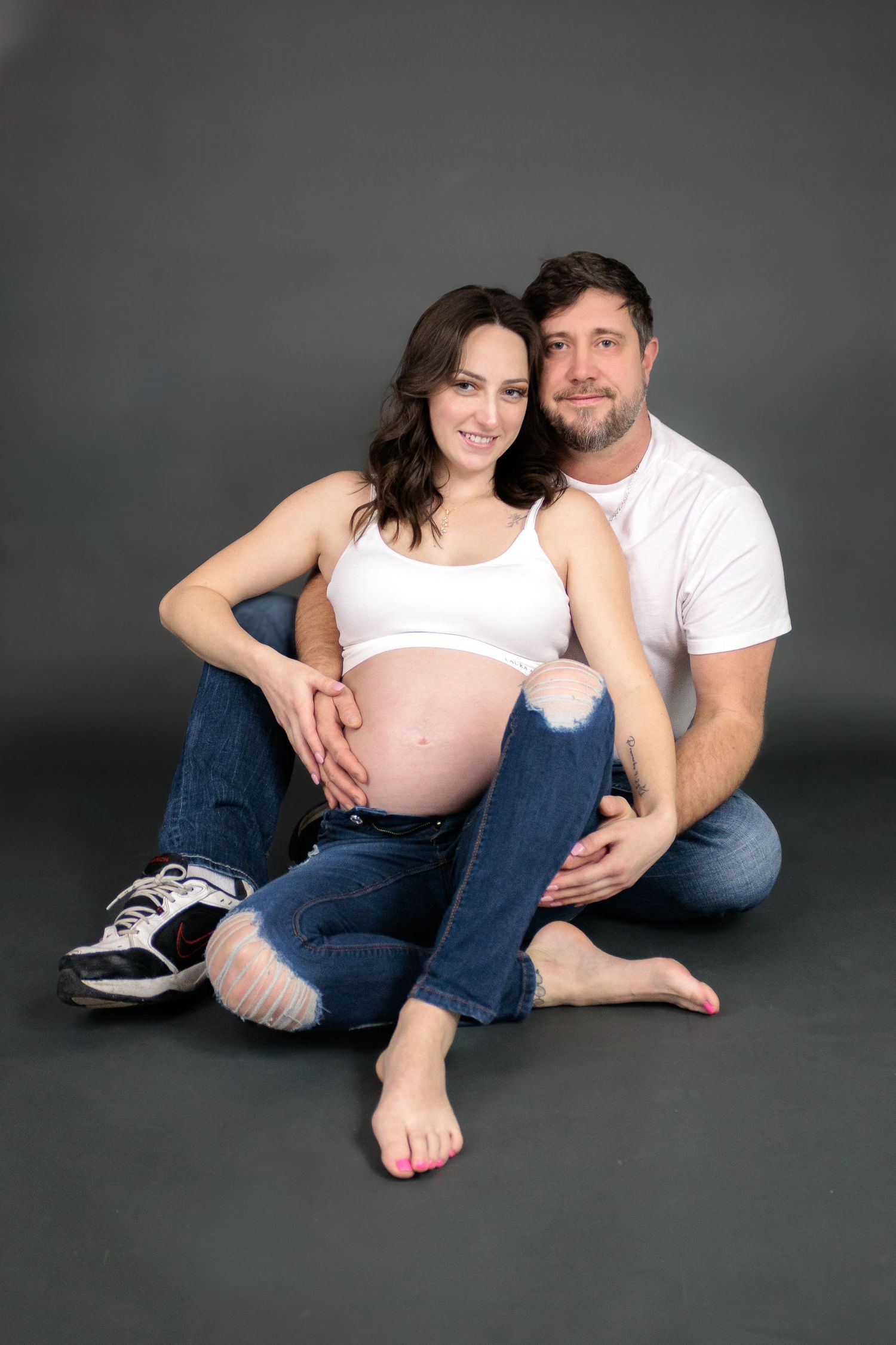 Natural maternity portraits of couple in casual wear on dark background.