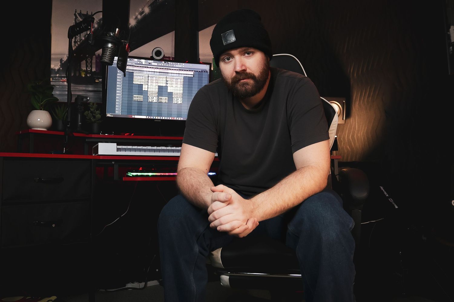 Music producer in black beanie sitting in recording studio with computer setup.