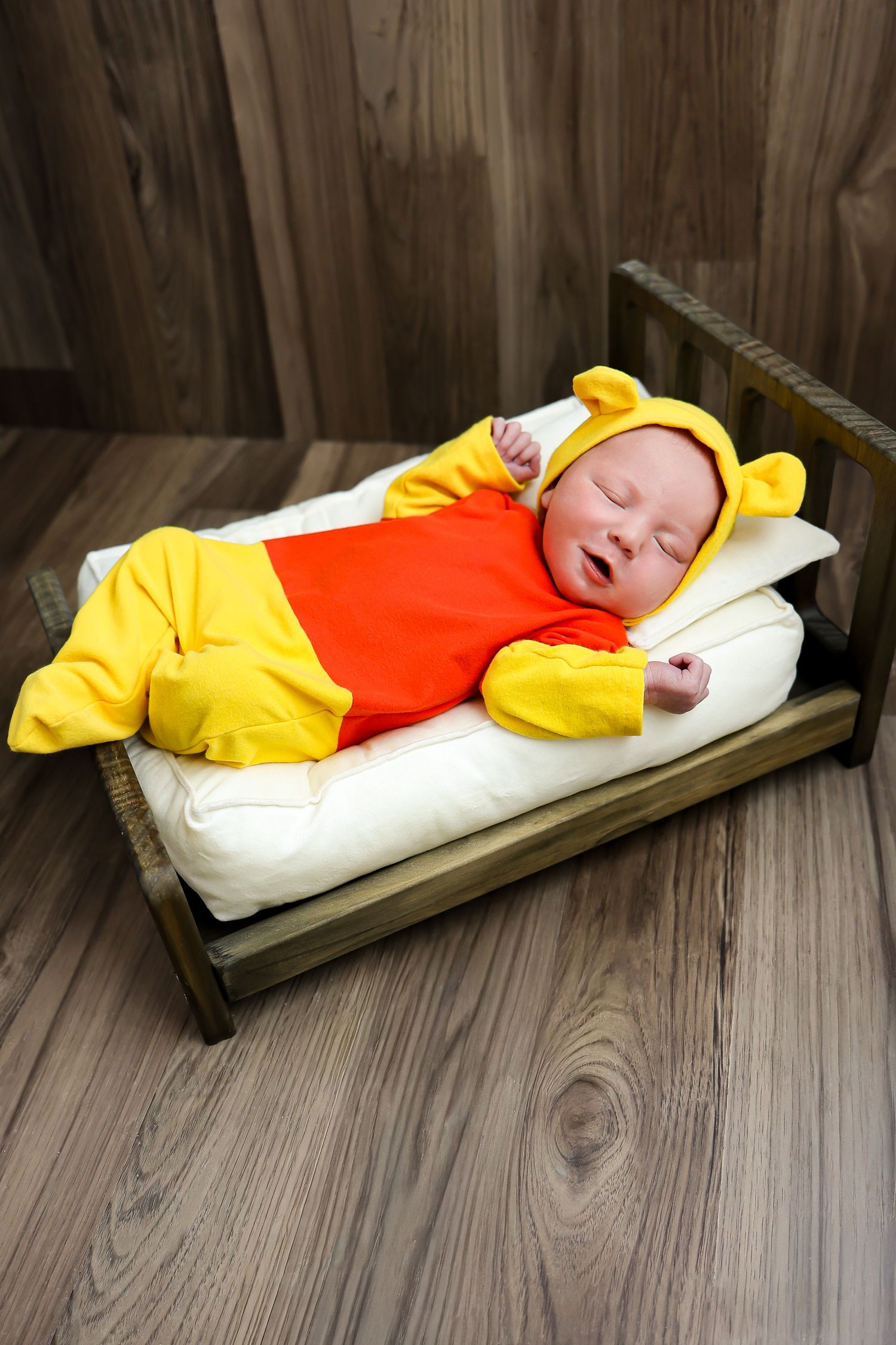 A baby dressed in a yellow and orange costume sleeps peacefully on a wooden swing.